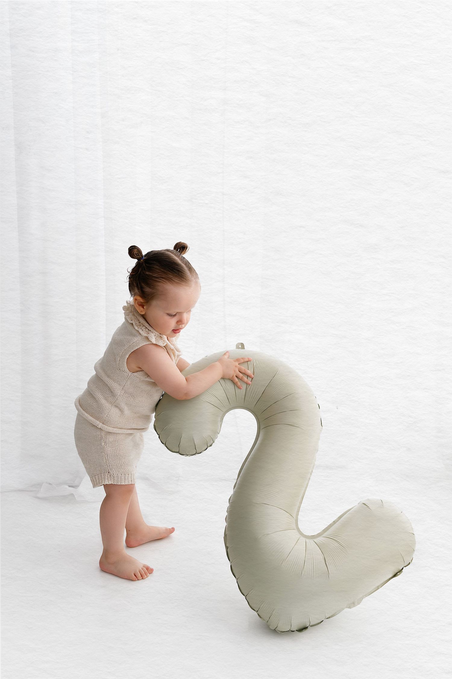Small child in light colored outfit playing with a large plush number 2 cushion on white background.