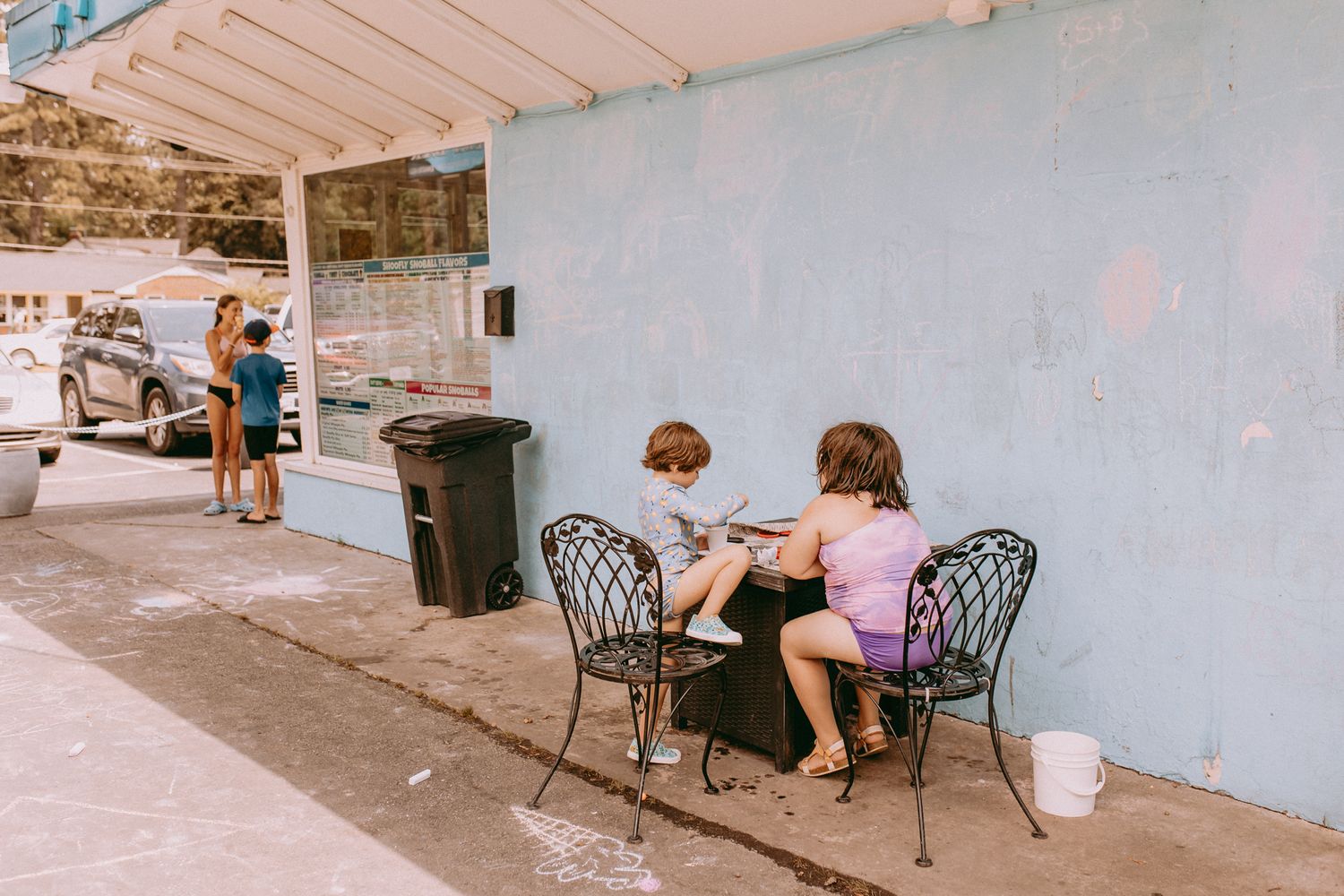 Two kids enjoy ice cream while sitting at bistro chairs against a blue wall with a walk up window visible in the background.