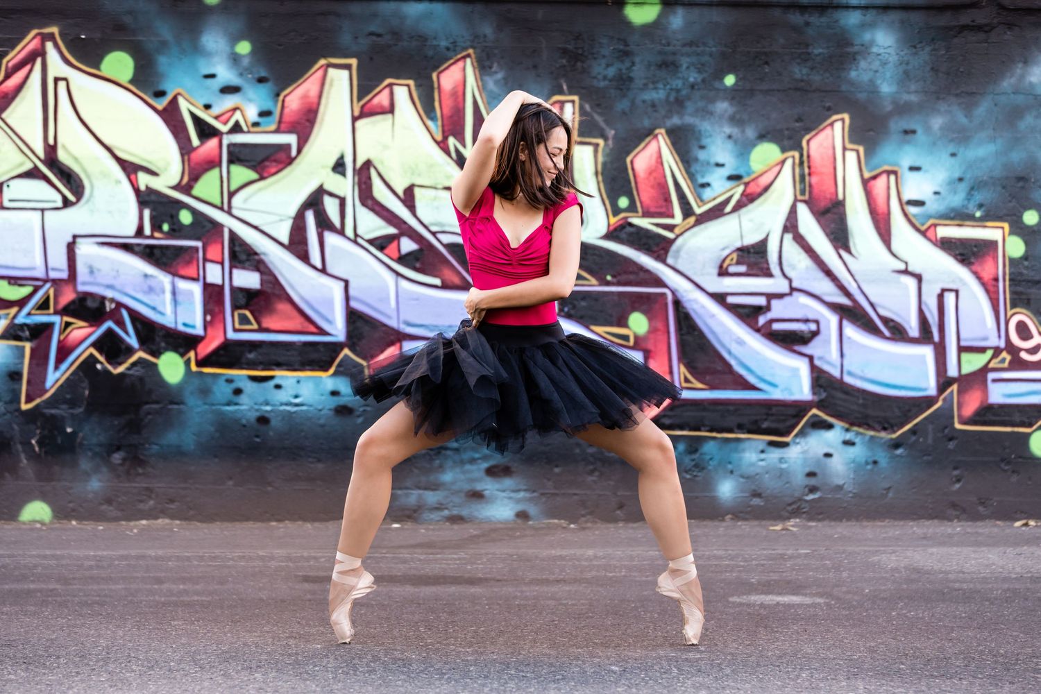 Dancer performs graceful ballet pose in front of vibrant graffiti wall wearing red top and black tutu.
