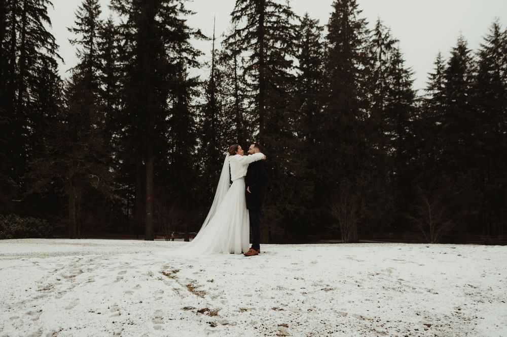 Winter wedding couple embracing in a snow-covered forest clearing with tall evergreen trees.