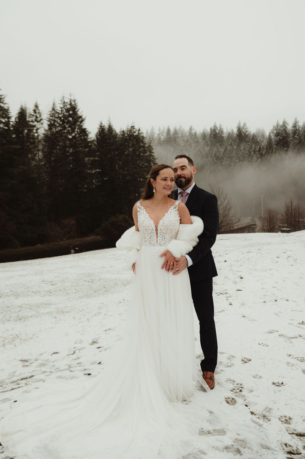 Elegant winter wedding portrait with couple standing in snow against evergreen forest backdrop.