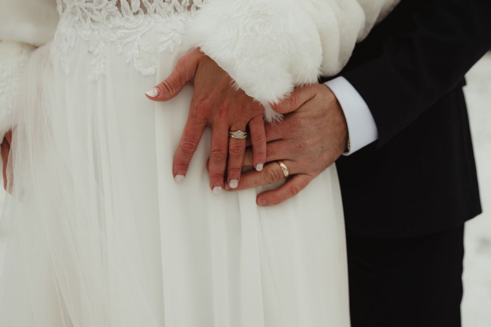 Close-up shot of hands wearing wedding rings against a white lace dress and black suit.