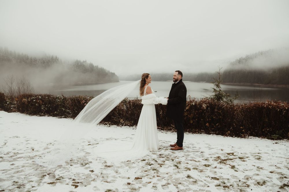 A dramatic winter wedding moment with a flowing veil caught in the wind against a foggy lake backdrop.