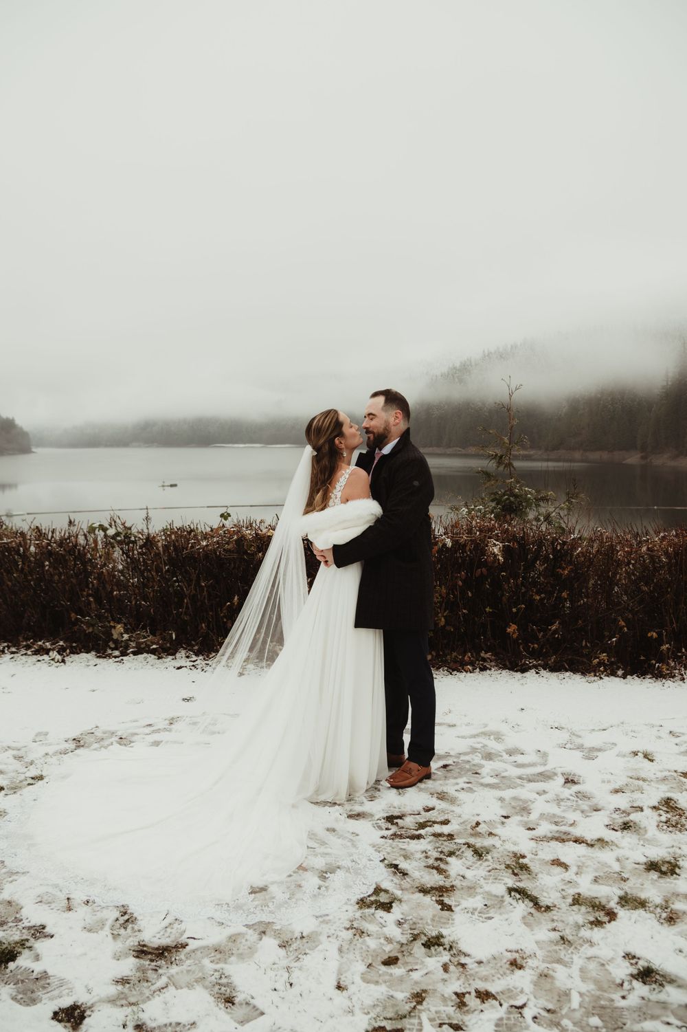 Romantic winter wedding portrait of a couple embracing in front of a misty lake landscape.