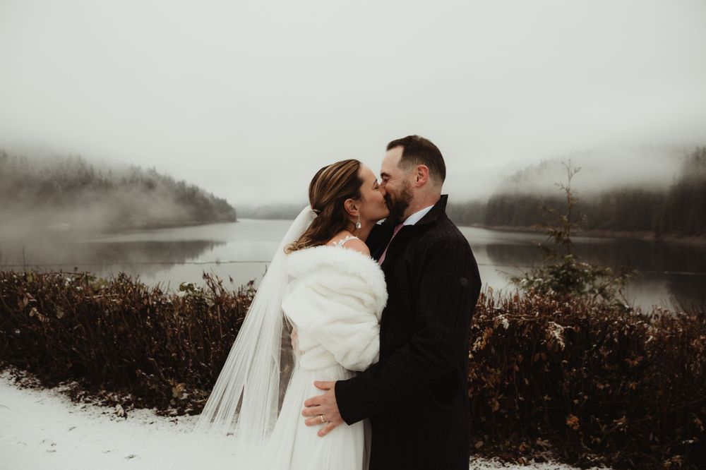 Intimate winter wedding moment captured against a moody lake backdrop with snow-covered ground.