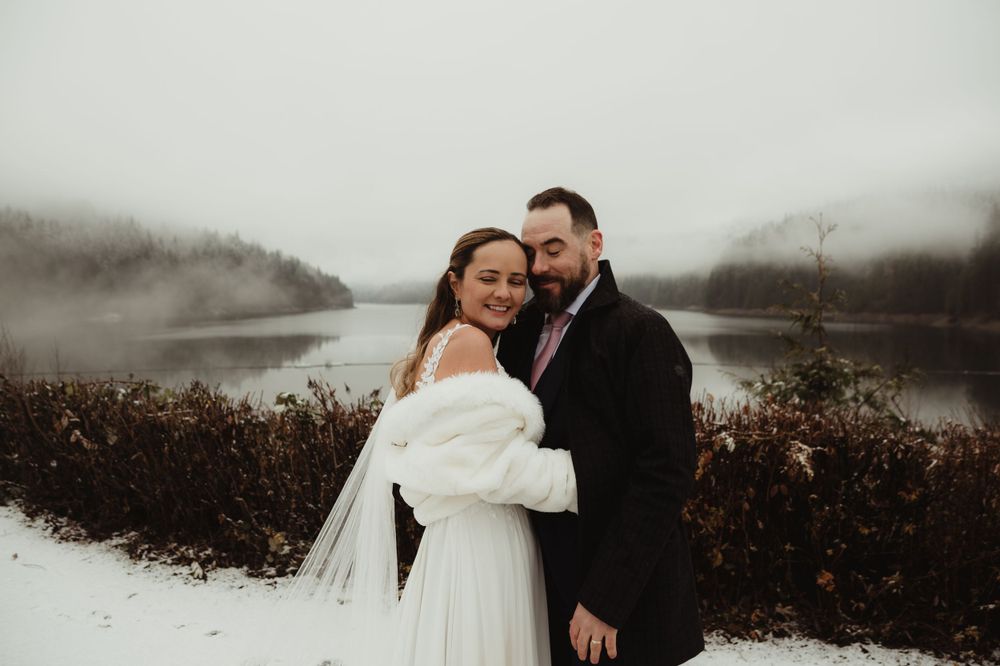 Happy newlyweds share a joyful moment in their winter wedding attire by a misty lake.