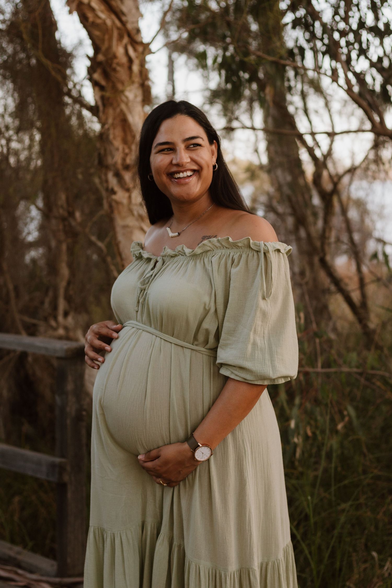 Pregnant woman wearing a green, flowy, off-the-shoulder dress. She's standing in a forest smiling and holding her belly