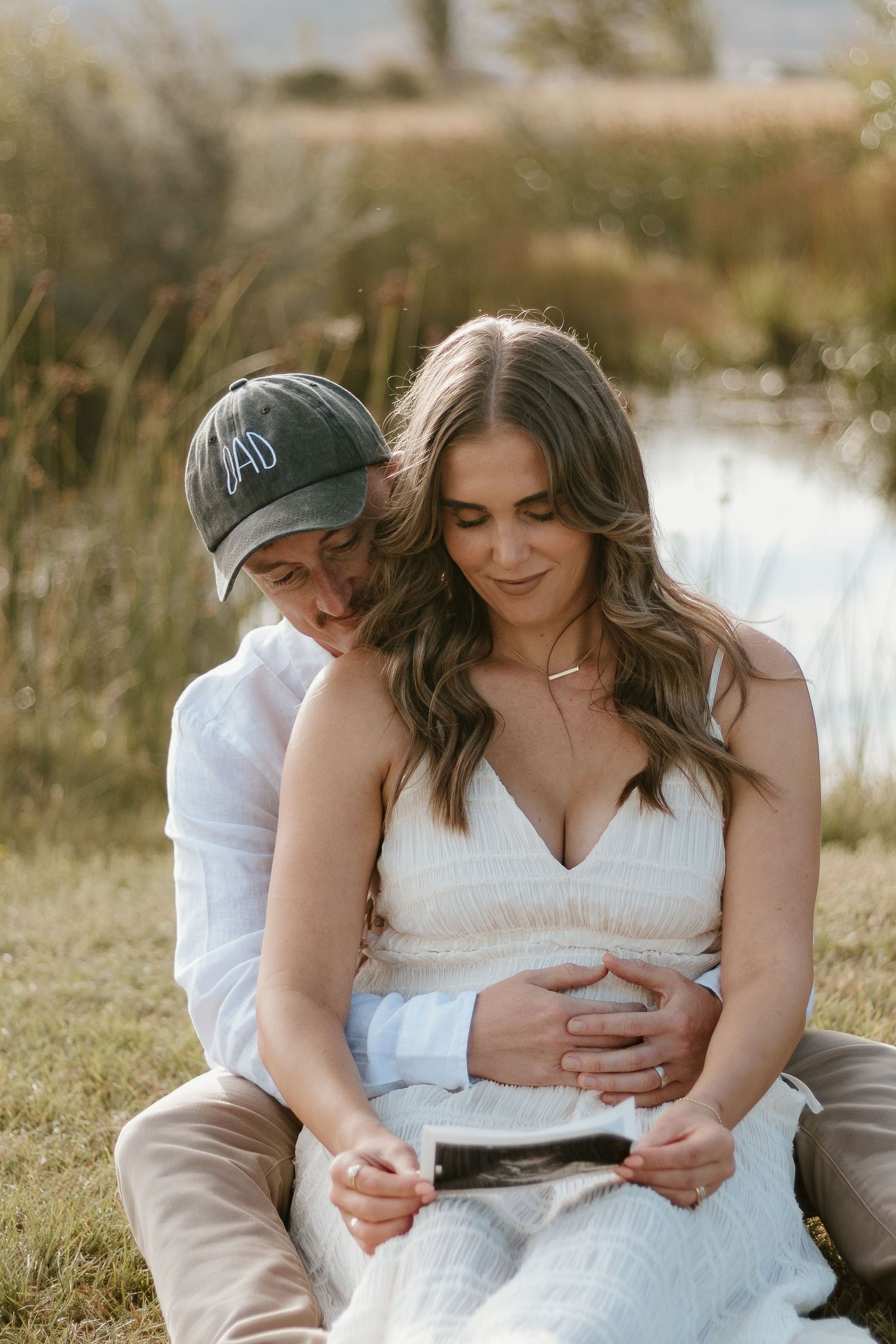 Expecting parents sitting in front of a pond, mum in between dad's legs with dad's arms around her waist. Looking at the sonogram