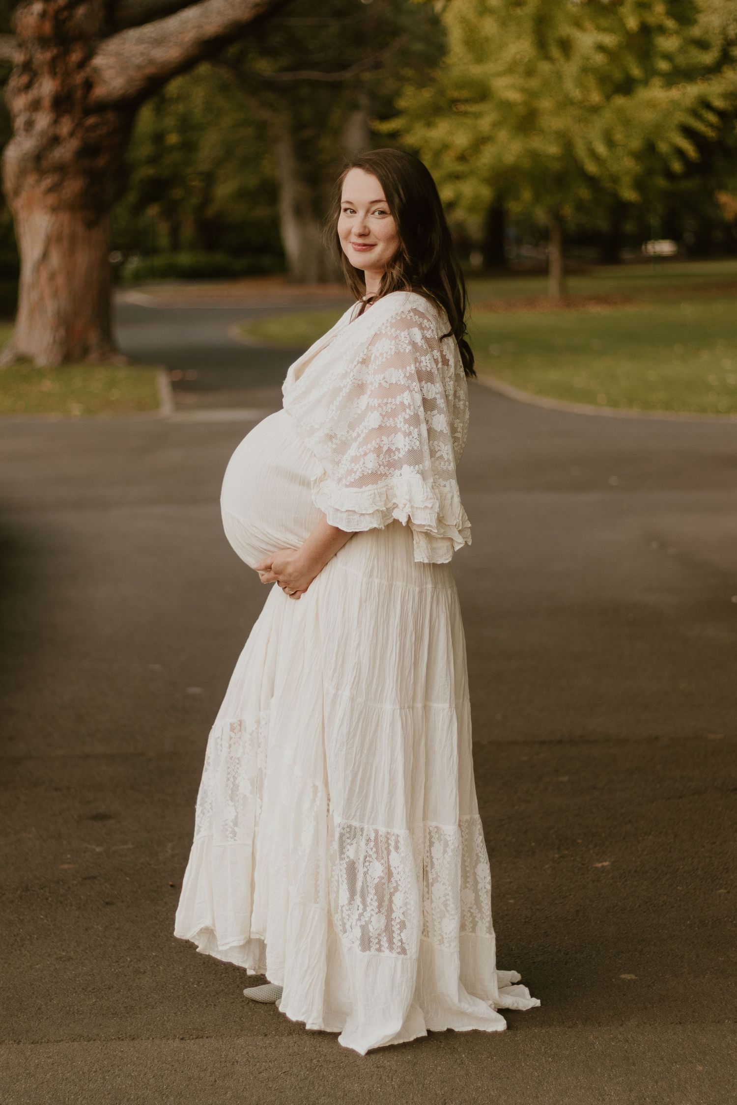 Pregnant woman wearing a long, flowy ivory dress, standing in City Park, Launceston, smiling and holding her belly
