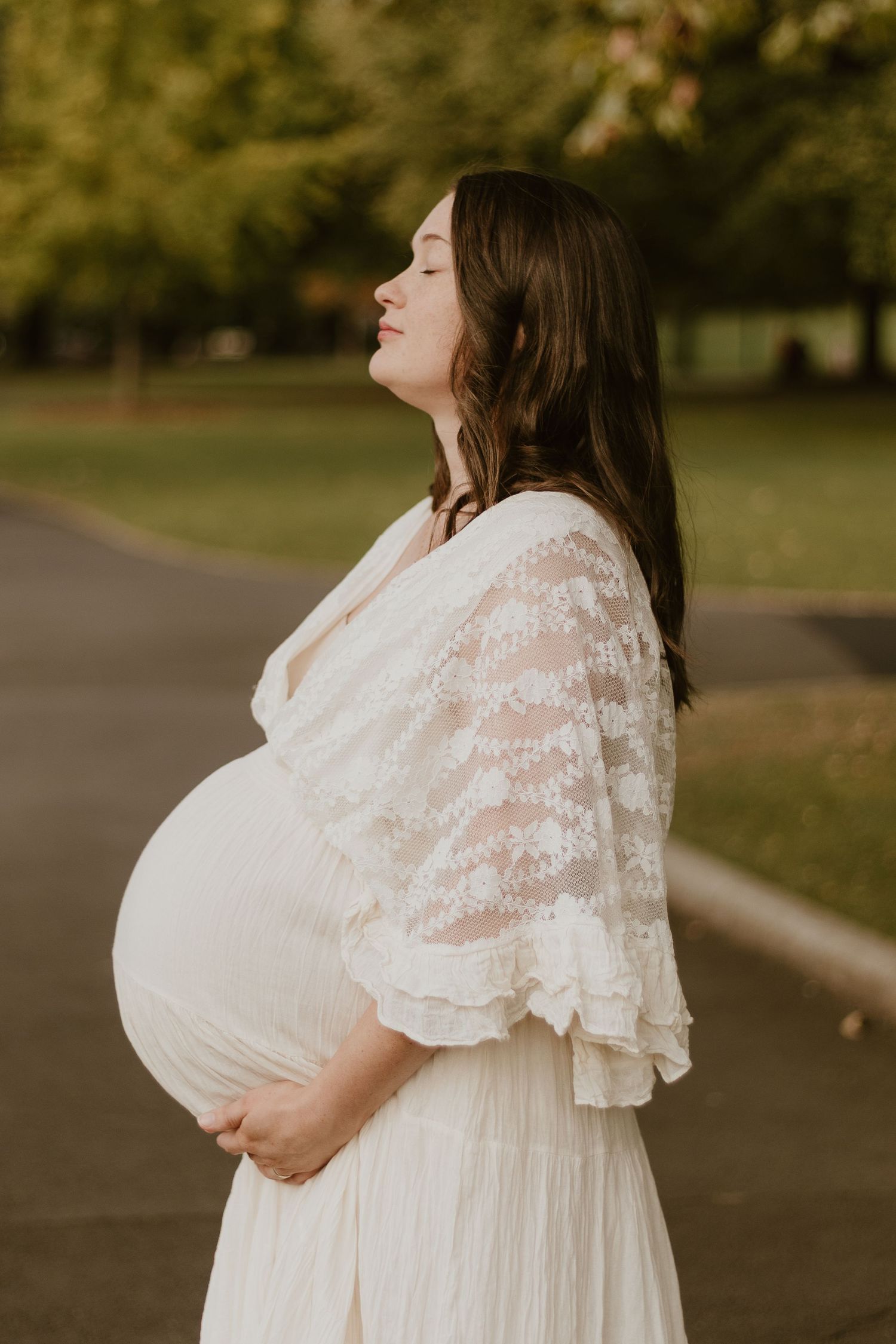 A pregnant woman standing in City Park, Launceston with her eyes closed and her hands underneath her pregnant belly