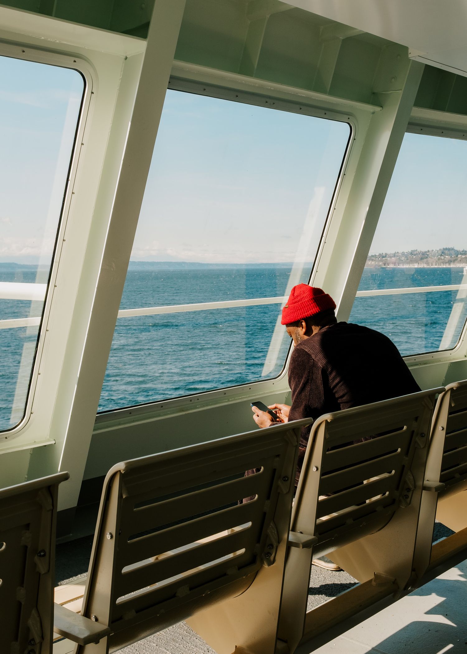 A passenger on a ferry wearing a red cap looks at their phone while sitting by large windows overlooking the ocean.