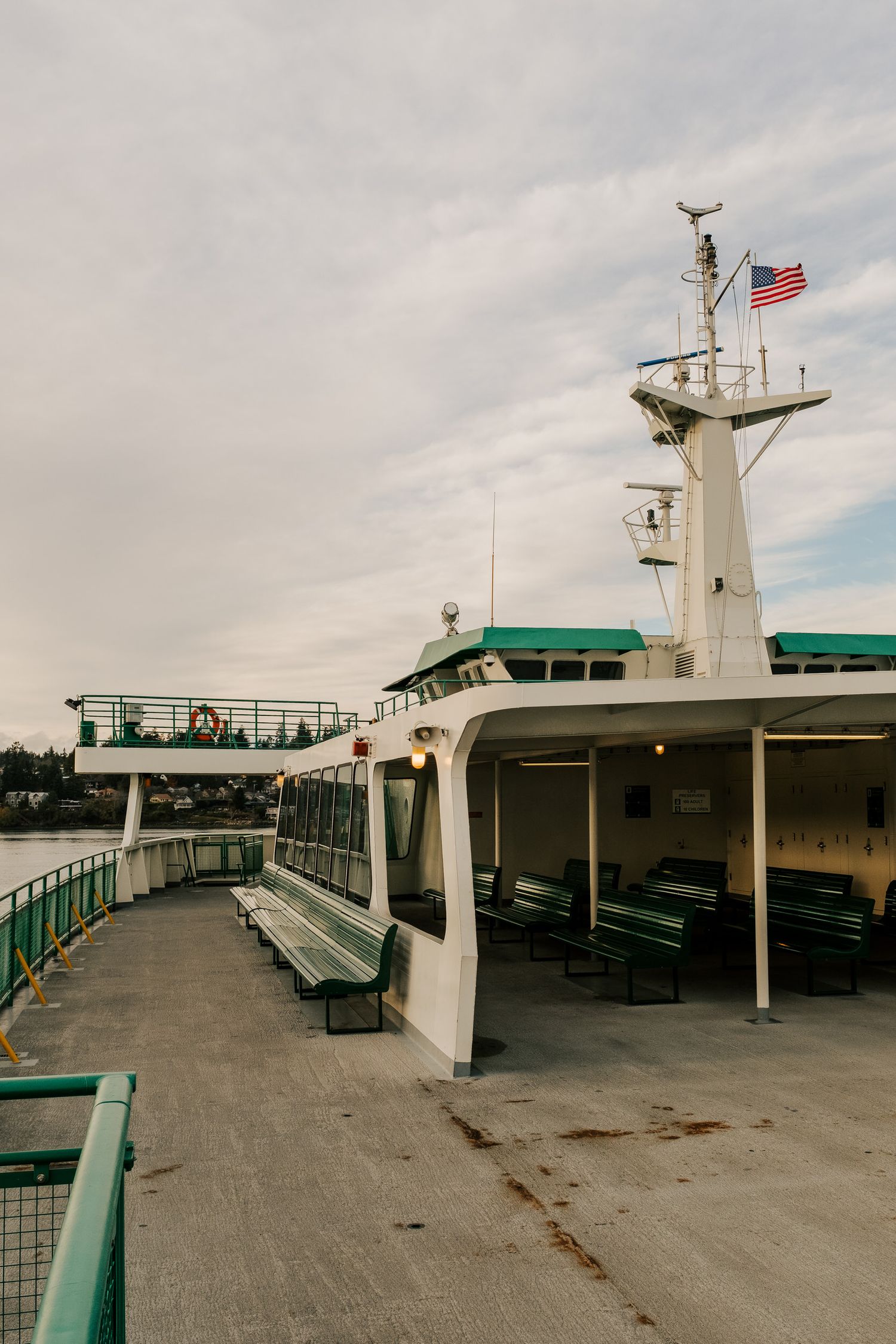 Ferry boat deck with benches and railings under cloudy sky with American flag flying from the mast.