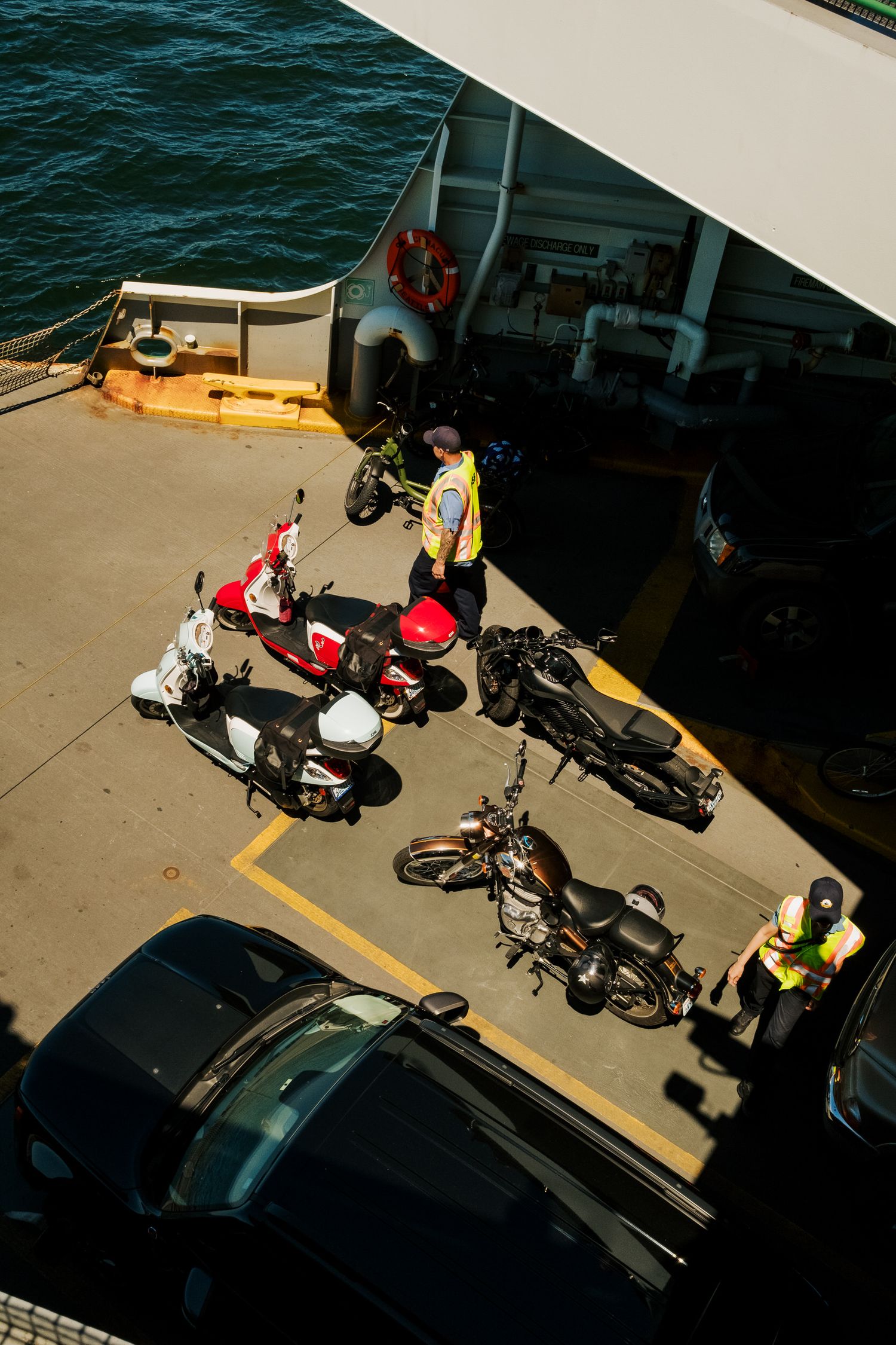 Motorcycles and bicycles parked on a ferry deck near deep blue water on a sunny day.