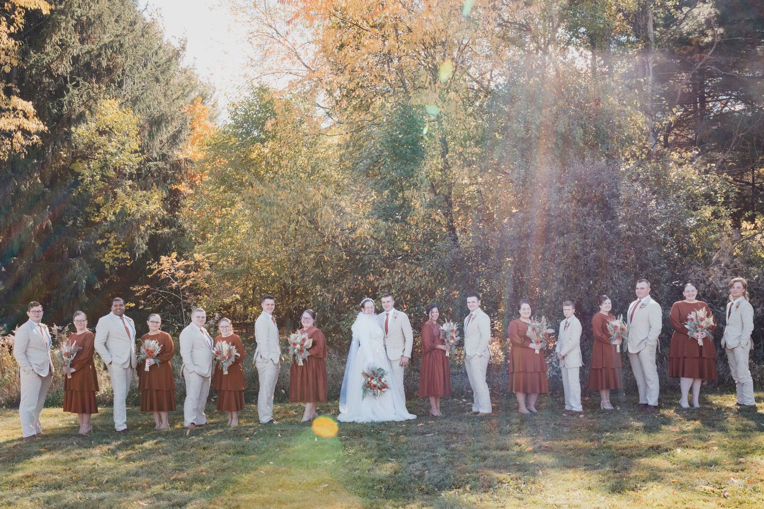 Wedding party lined up for group photos in autumn outdoor setting with trees.