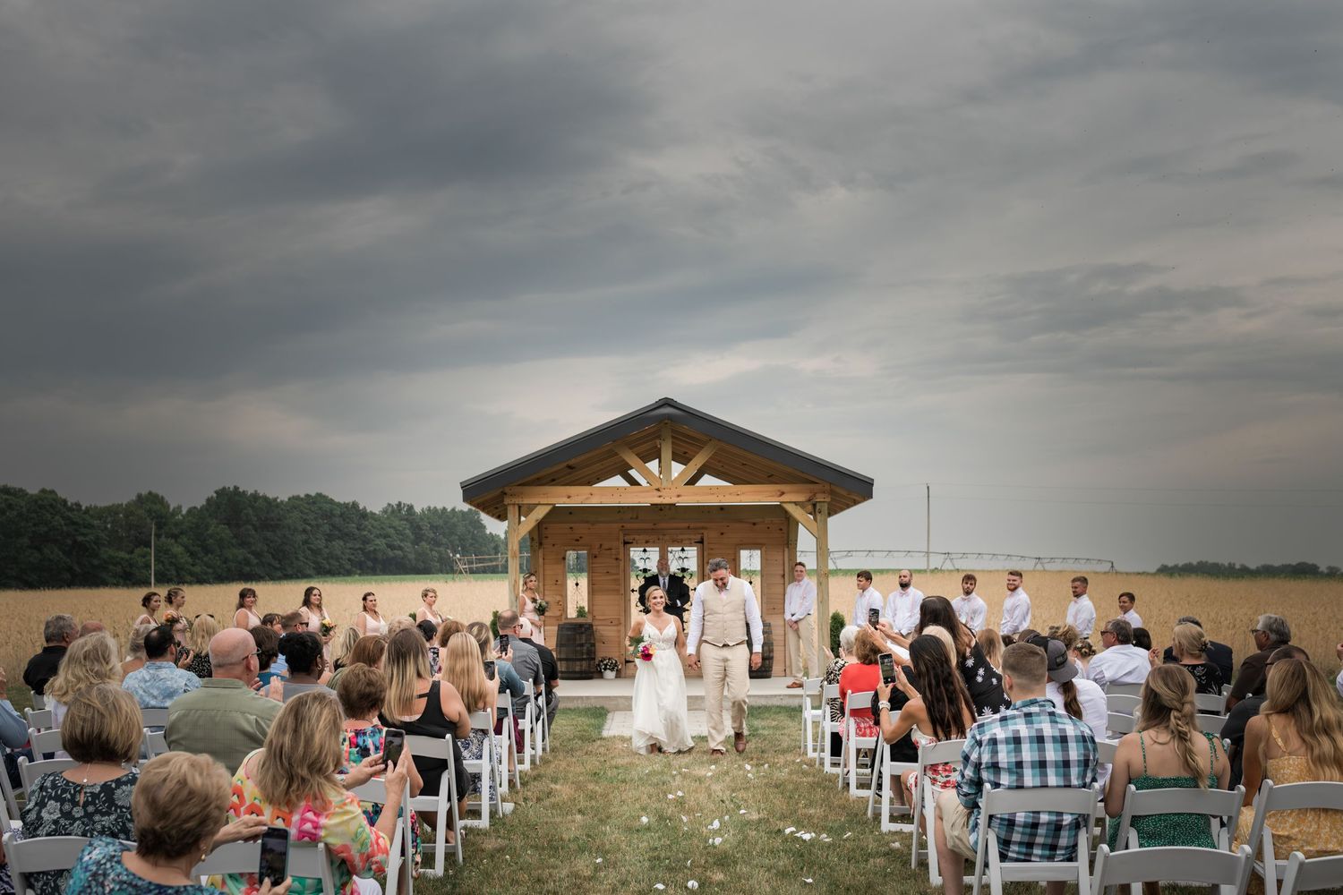 Outdoor wedding ceremony setup with wooden arch and white chairs under cloudy sky.