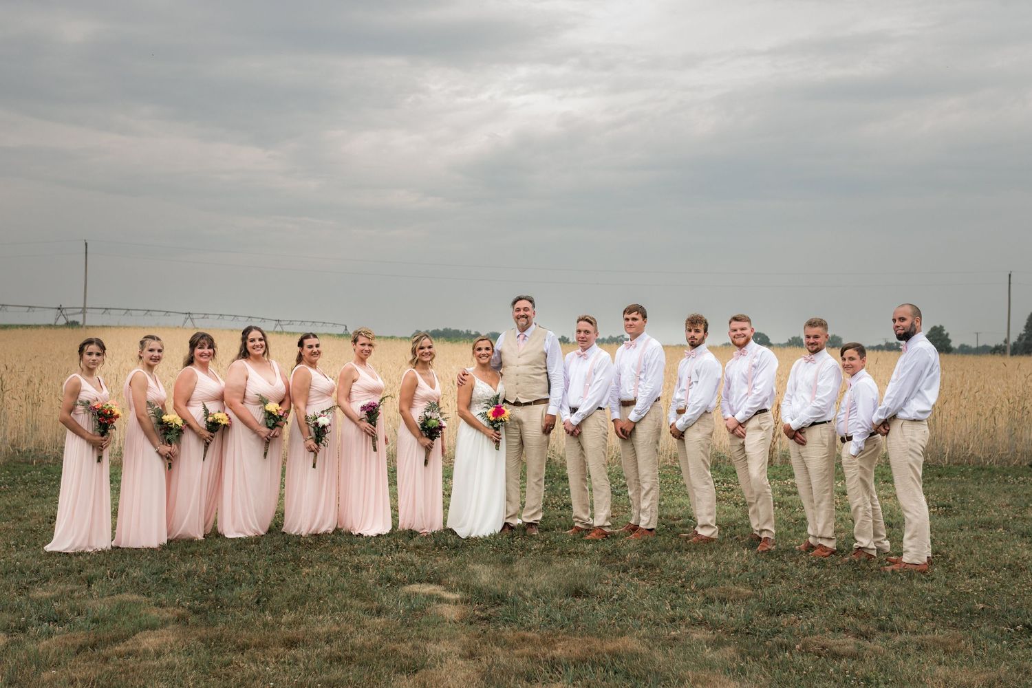 Wedding party lined up in blush and white attire for panoramic group photo in field.