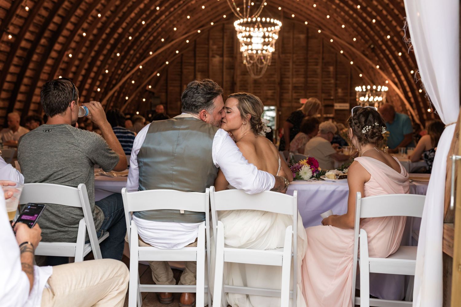 Wedding reception guests seated in white chairs under elegant chandeliers in a rustic barn venue.