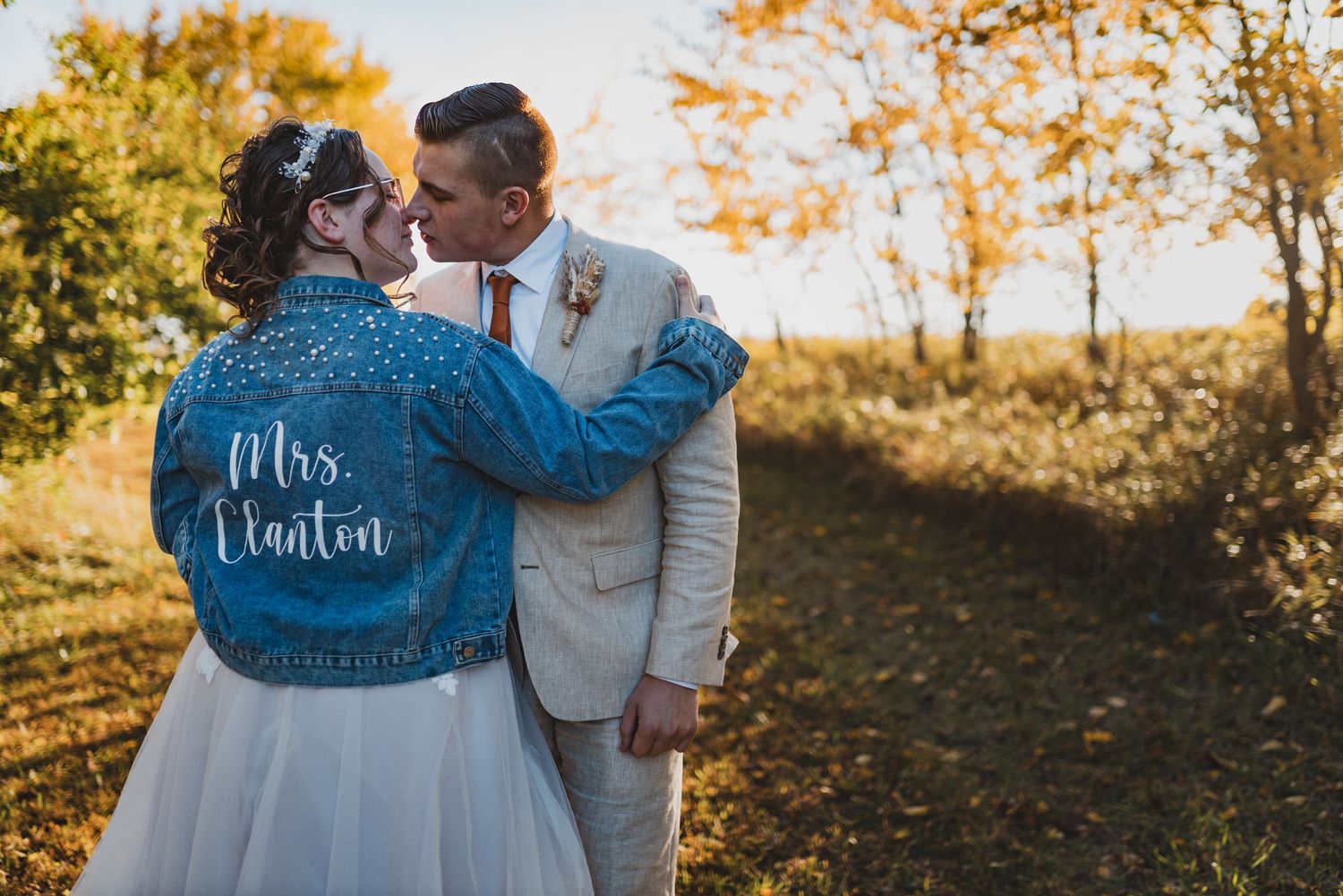 Couple wearing custom denim jackets with wedding titles embraces in golden sunlight.