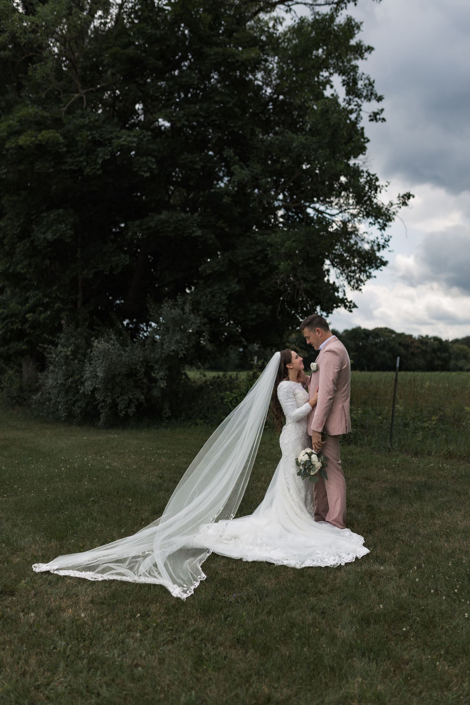 Dramatic wedding photos in field with flowing white dress train against cloudy sky.