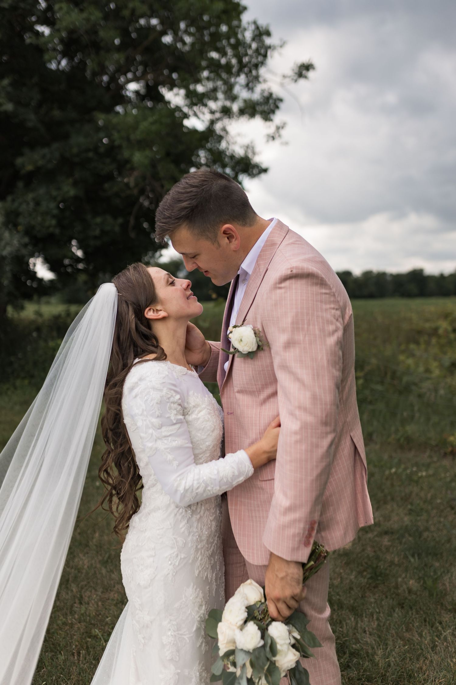 Couple in pink suit and white lace dress share intimate moment in outdoor field setting.
