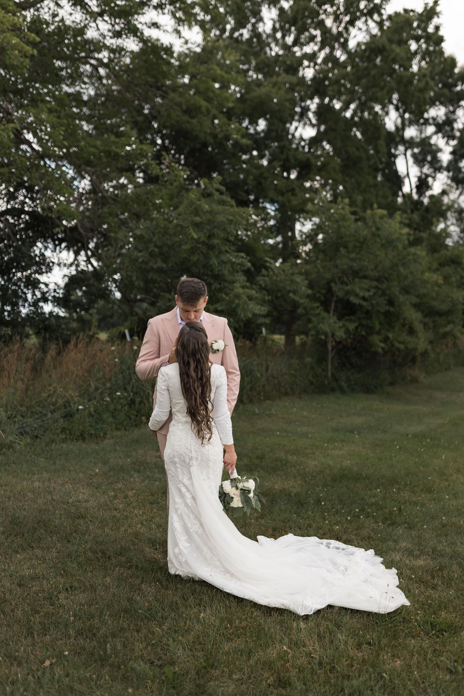 Elegant wedding couple poses in grassy field with dramatic train of white dress.