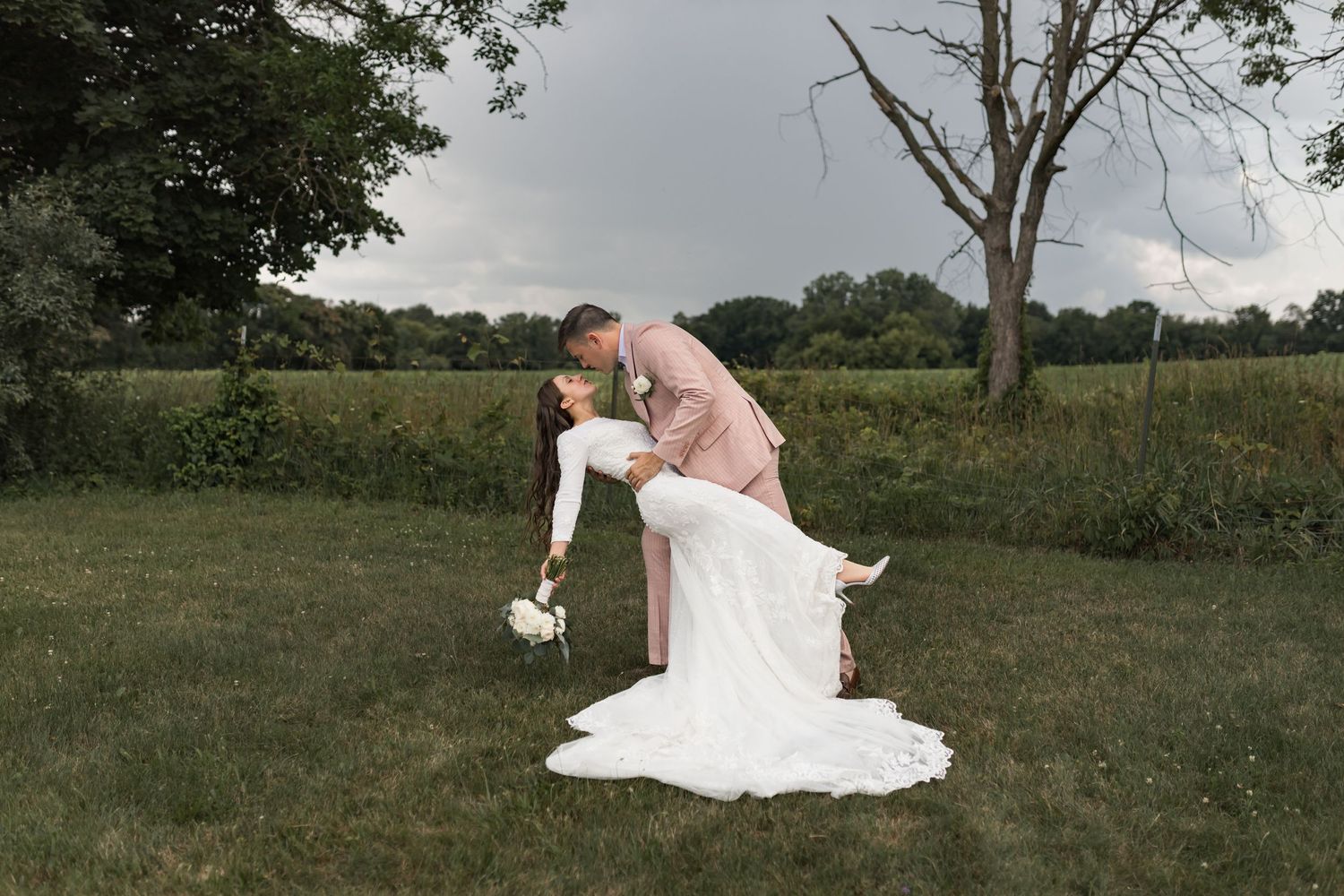 Wedding couple embraces in outdoor field setting with dramatic dress train.