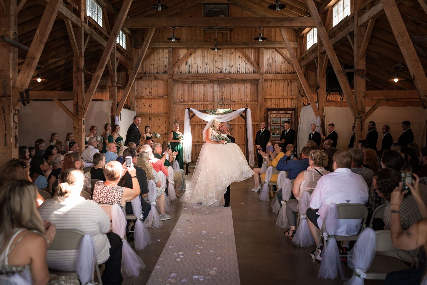 Wedding ceremony setup inside rustic wooden barn with rows of white chairs.