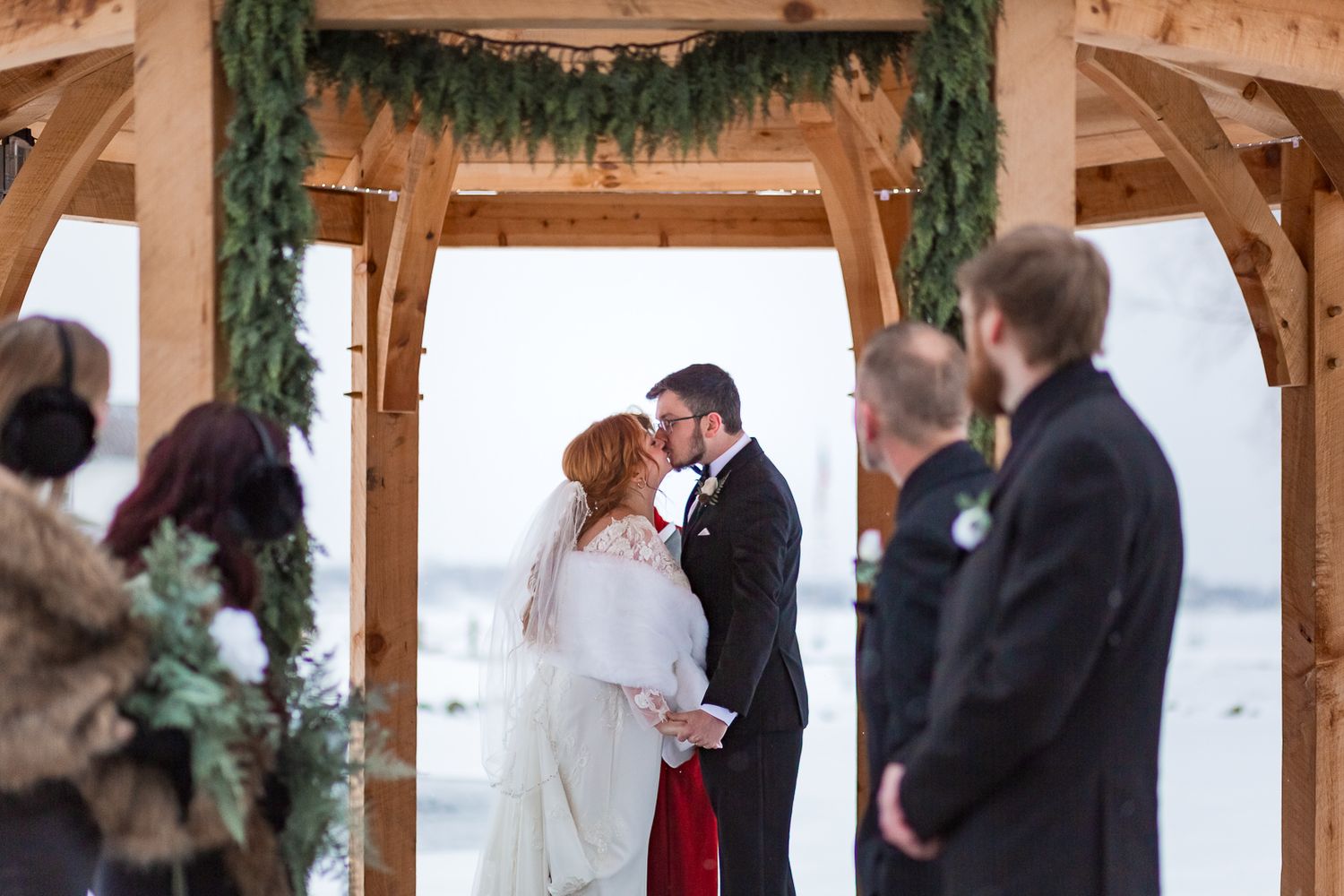 Winter wedding ceremony under a snow-covered wooden gazebo with guests seated outdoors.