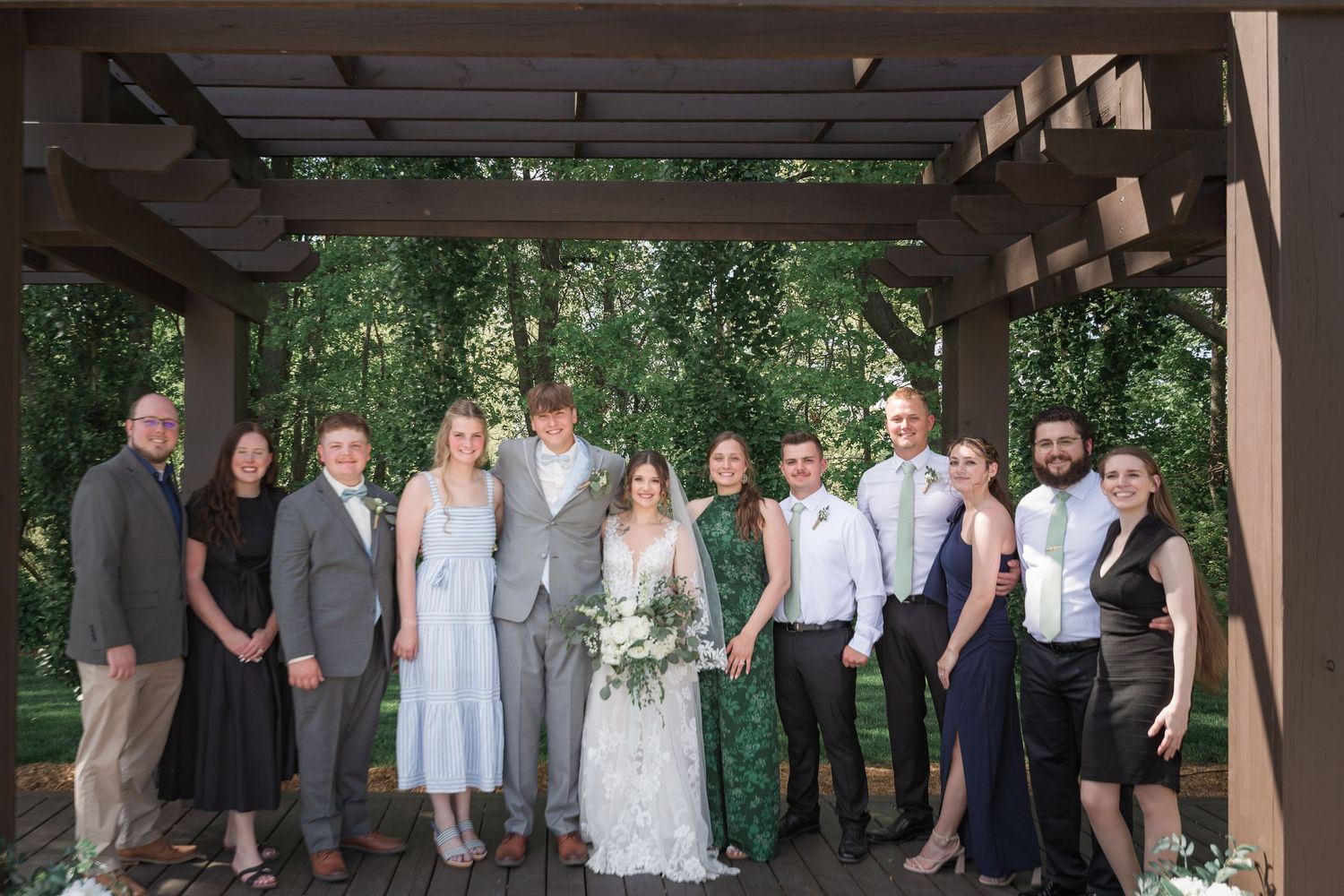 Large wedding party group photo taken under covered outdoor pavilion.