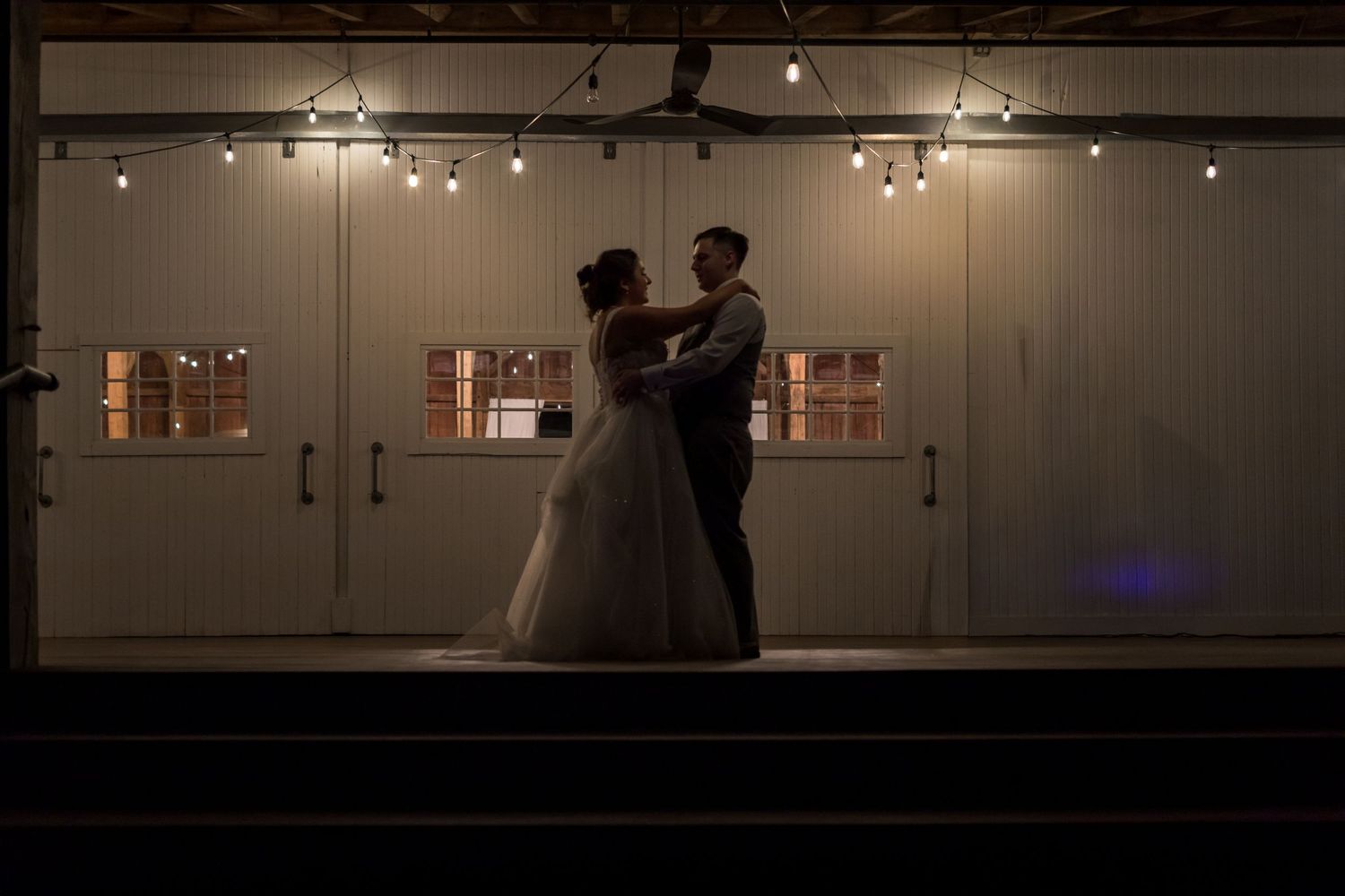 Silhouetted bride and groom share first dance in dimly lit barn venue with string lights glowing overhead.
