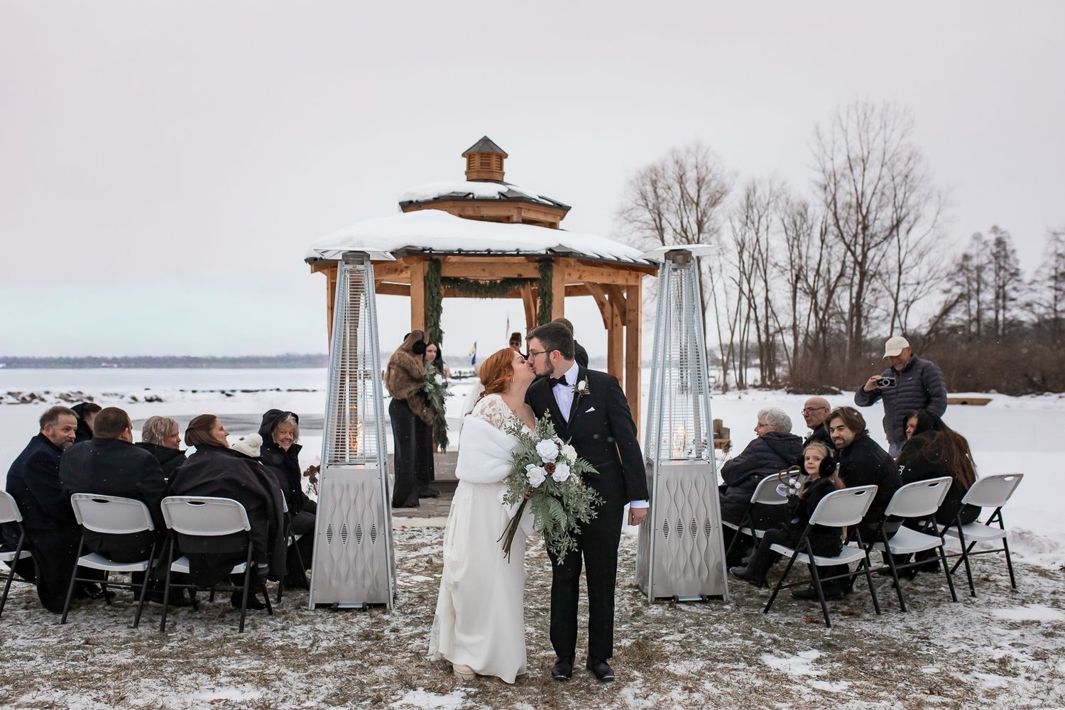 Winter wedding ceremony setup with white chairs on snow-covered ground beneath a wooden pergola.