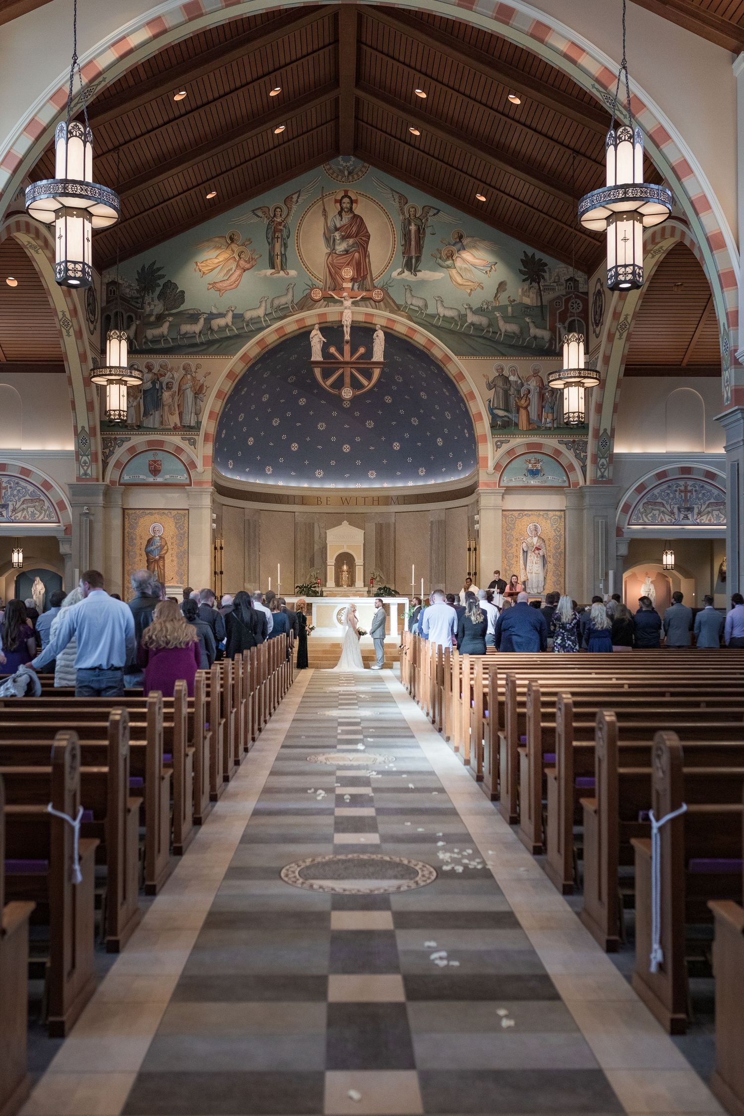 Interior of a grand church with wooden pews and ornate ceiling murals illuminated by hanging pendant lights.