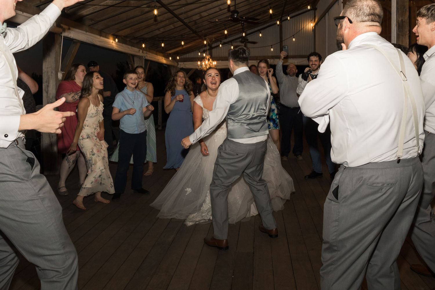 Bride and groom dancing joyfully with wedding guests on a rustic wooden dance floor at their reception.