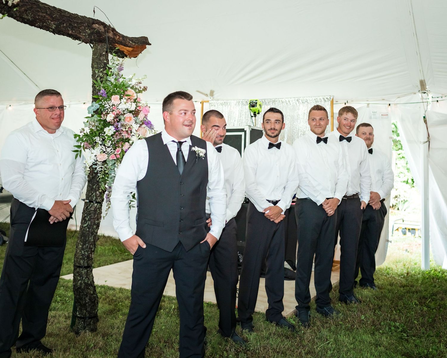 Wedding party lined up outdoors wearing formal black and white attire near a white tent.
