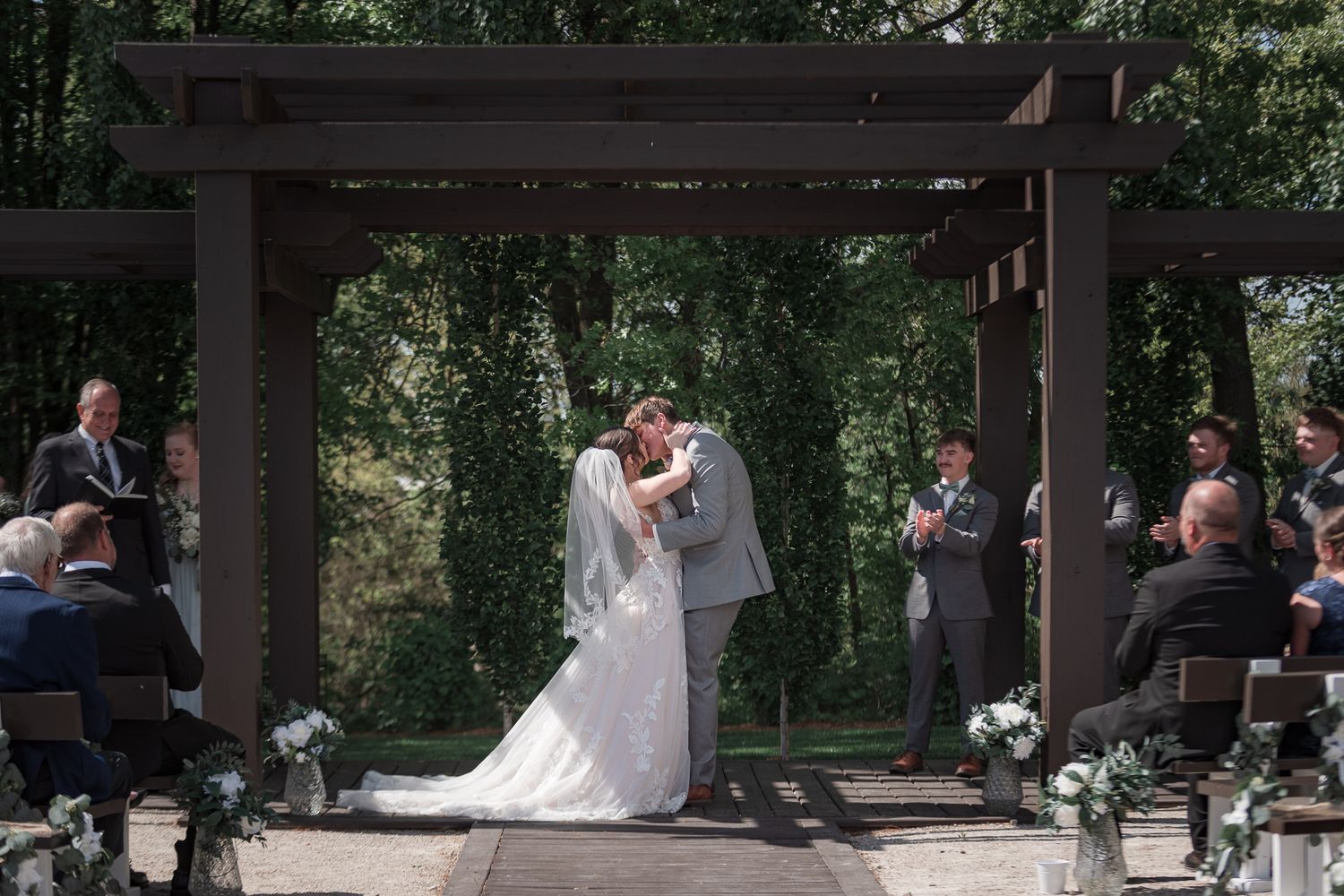 A wedding ceremony takes place under a rustic wooden pergola surrounded by lush greenery and guests.