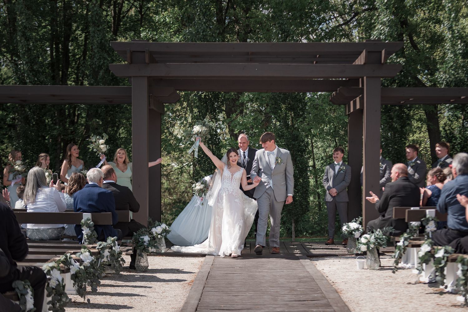 Outdoor wedding ceremony scene with guests seated beneath a wooden pavilion in a garden setting.