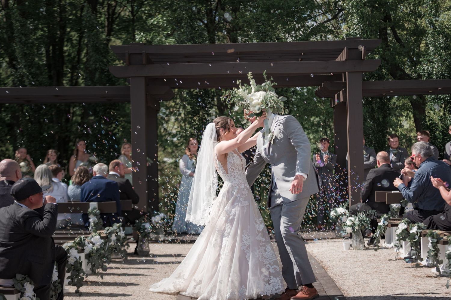 Wedding celebration under a wooden gazebo with guests watching from wooden benches.