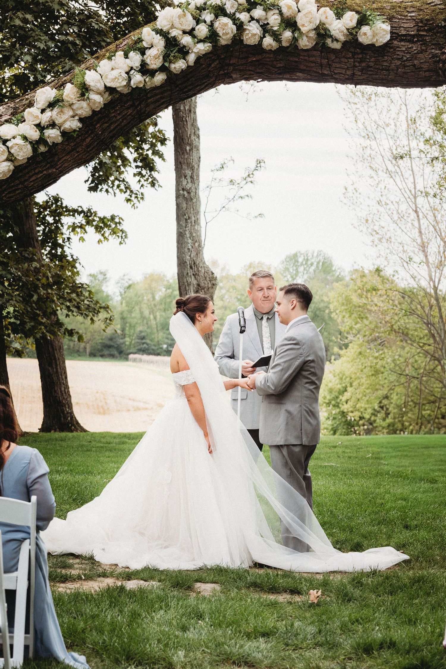 Bride and groom exchange vows outdoors under a floral arch with white roses, officiant holding book, lush green backdrop.