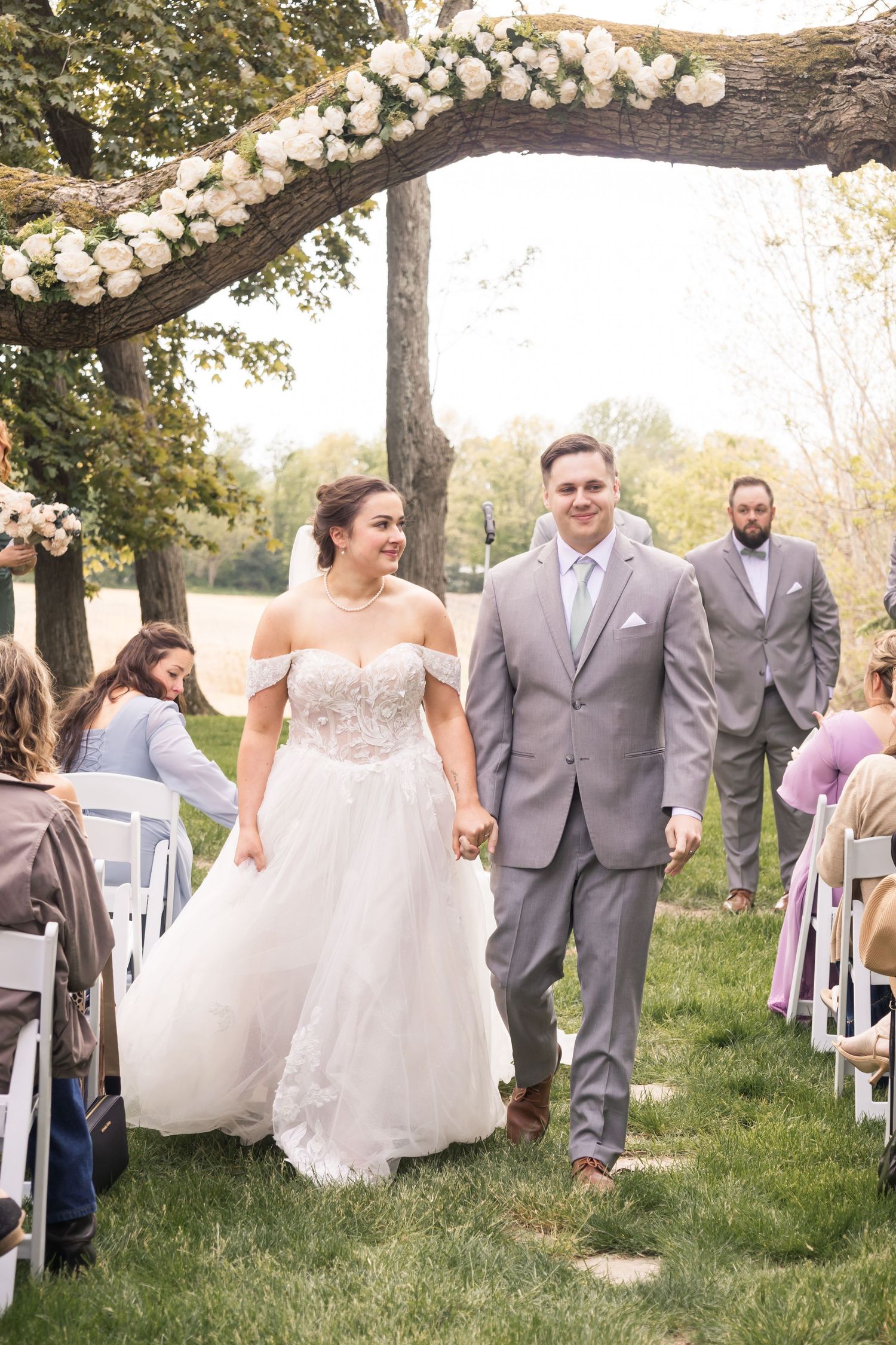 Bride and groom walk down outdoor wedding aisle under floral arch, smiling joyfully, surrounded by seated guests on a sunny day.