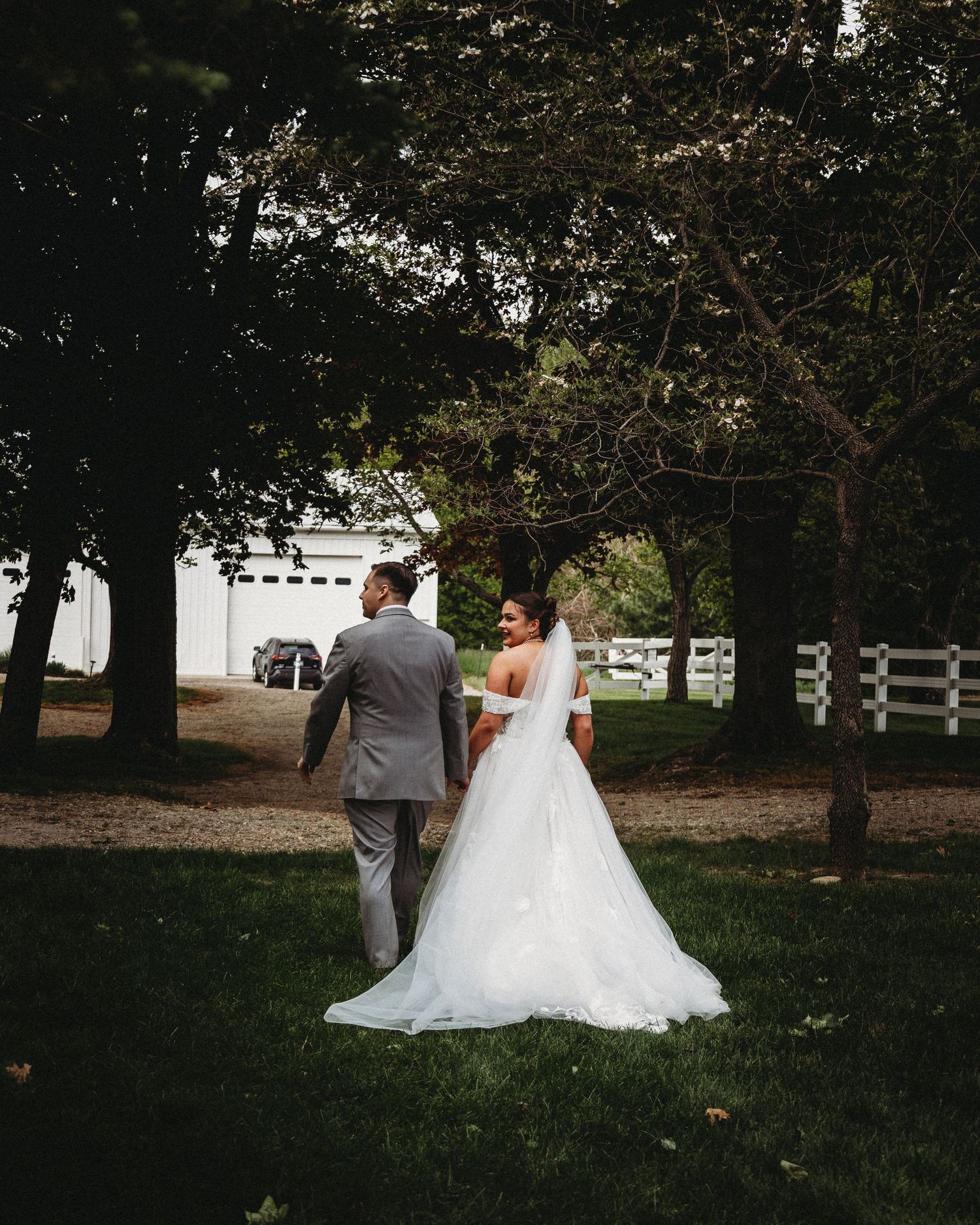 Bride in white gown with veil and groom in gray suit walking through tree-lined lawn near white barn.