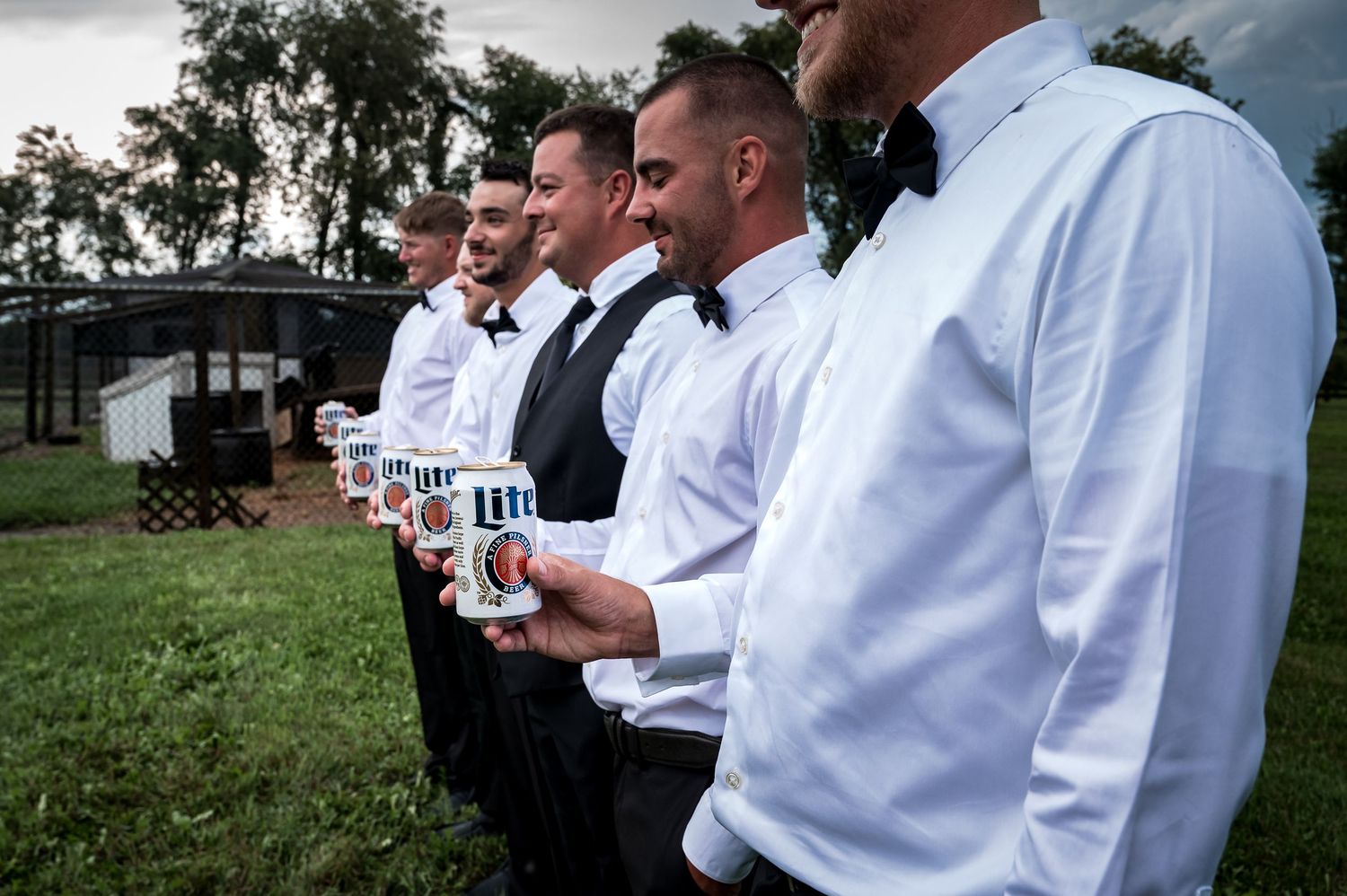 Groomsmen in white shirts and bow ties toasting with drinks outdoors.