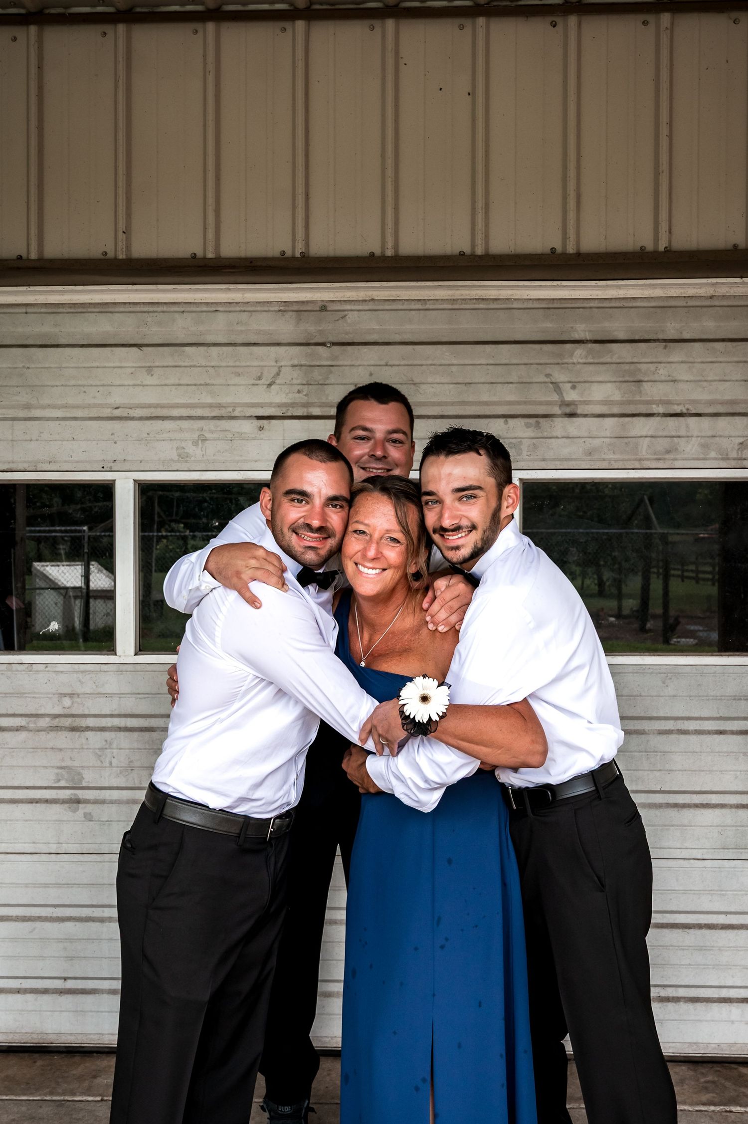 Wedding party members in formal attire share a group hug against a white building.