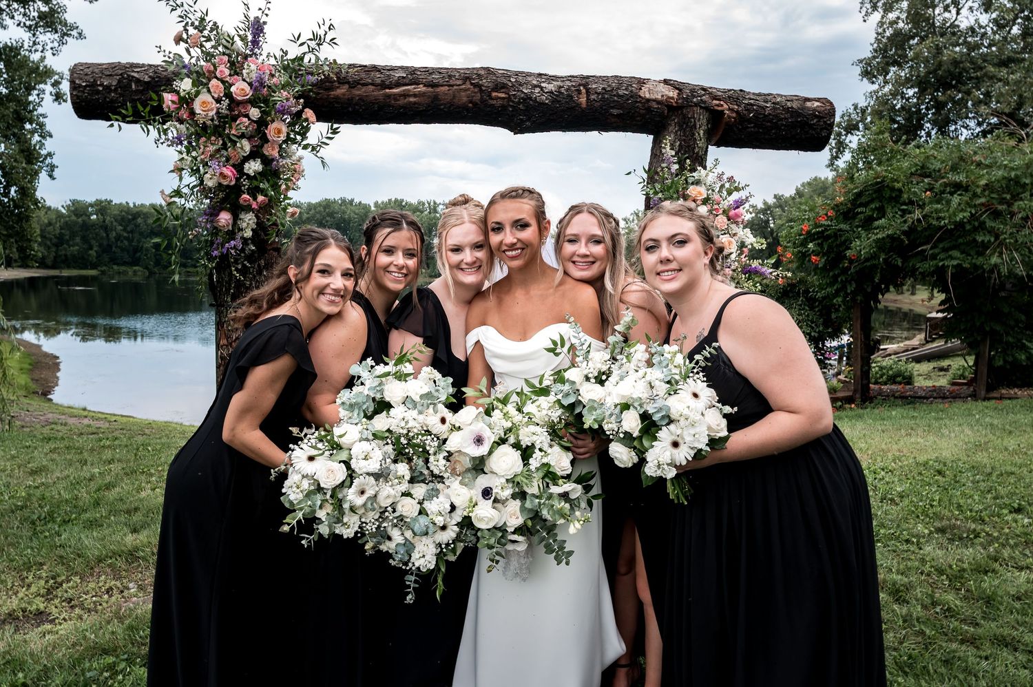 Bridesmaids in elegant dresses pose with white floral bouquets against a rustic wooden backdrop.