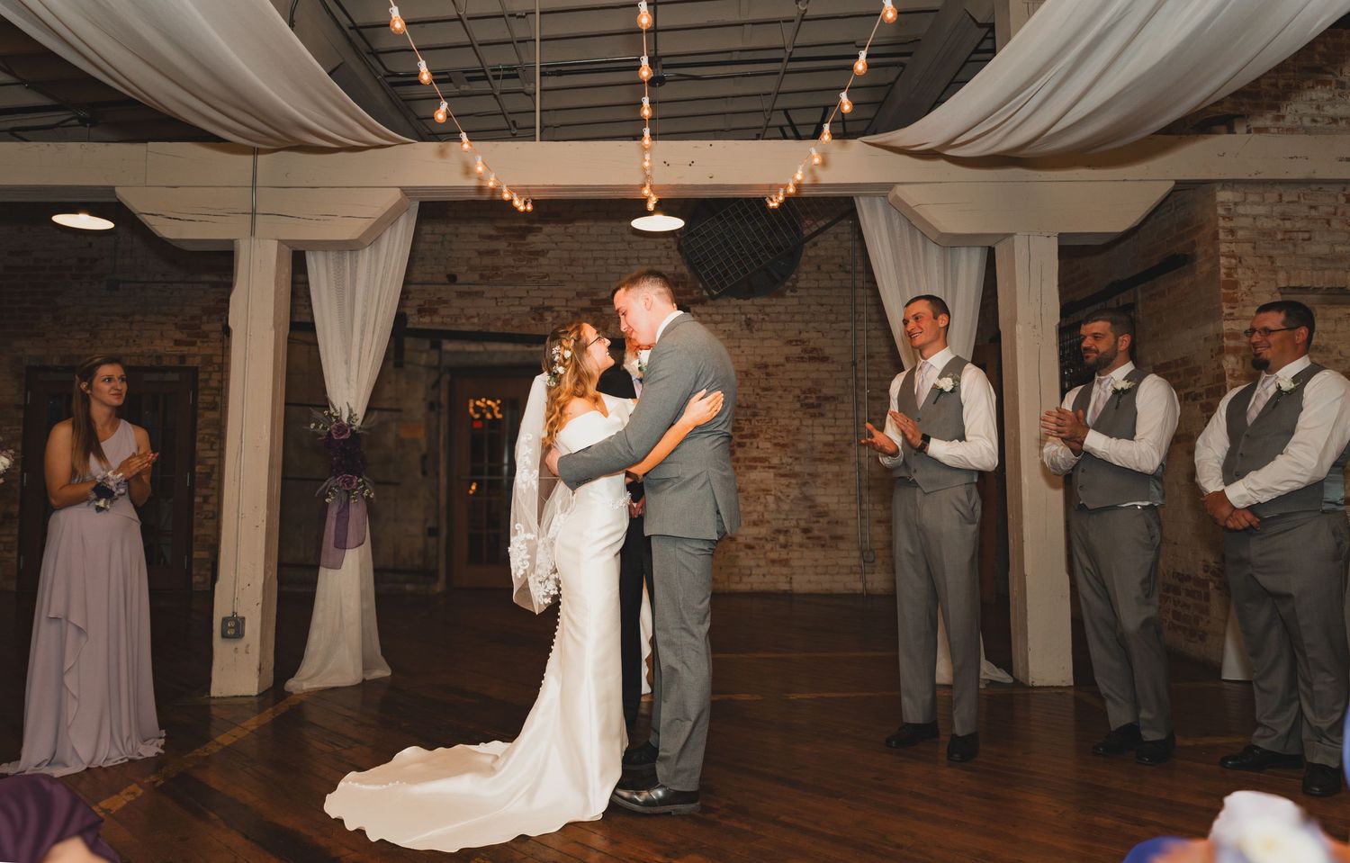Newlyweds share their first dance surrounded by wedding party in rustic reception venue.
