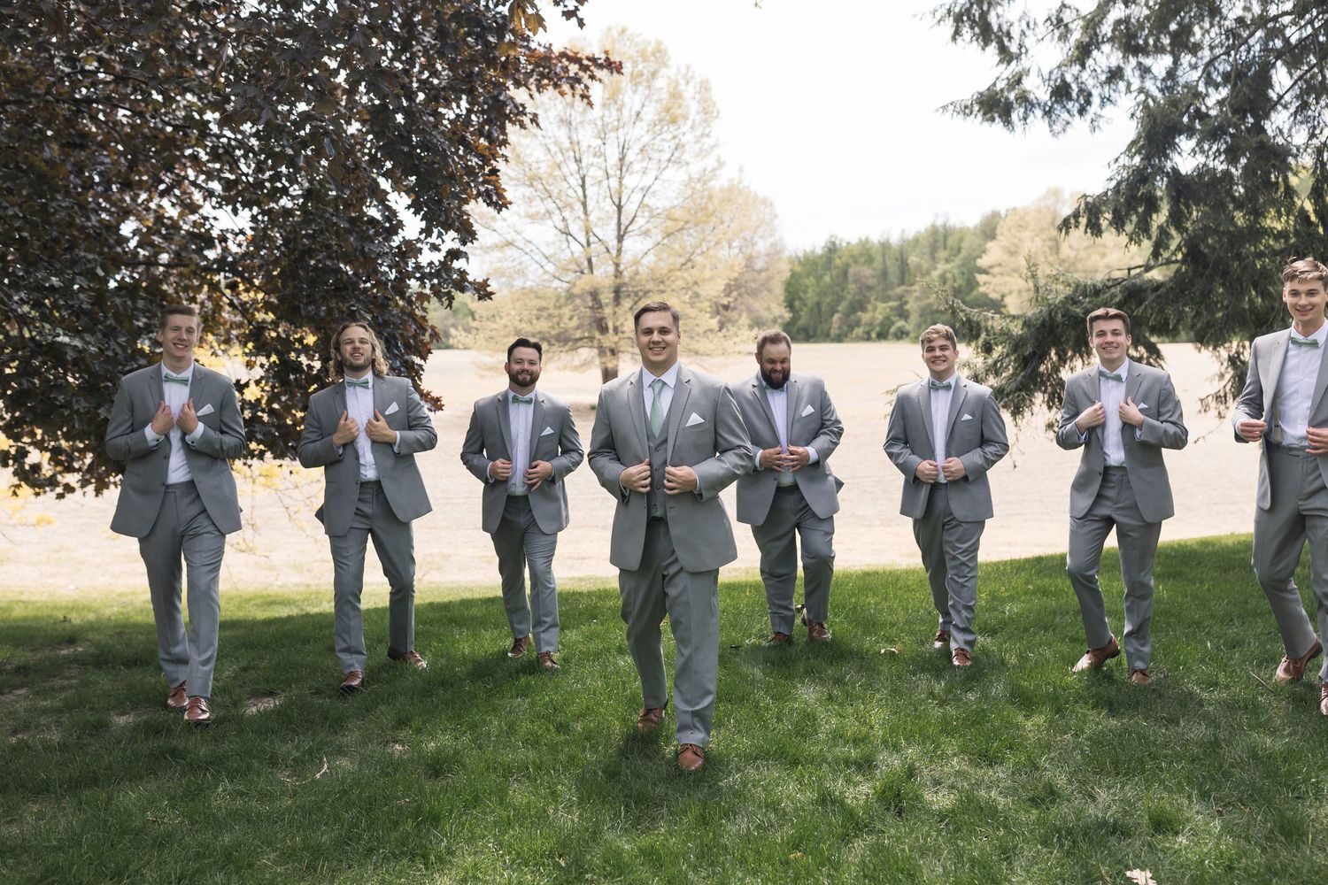 Groom leads groomsmen in matching gray suits and pink bow ties on a green lawn at an outdoor wedding.