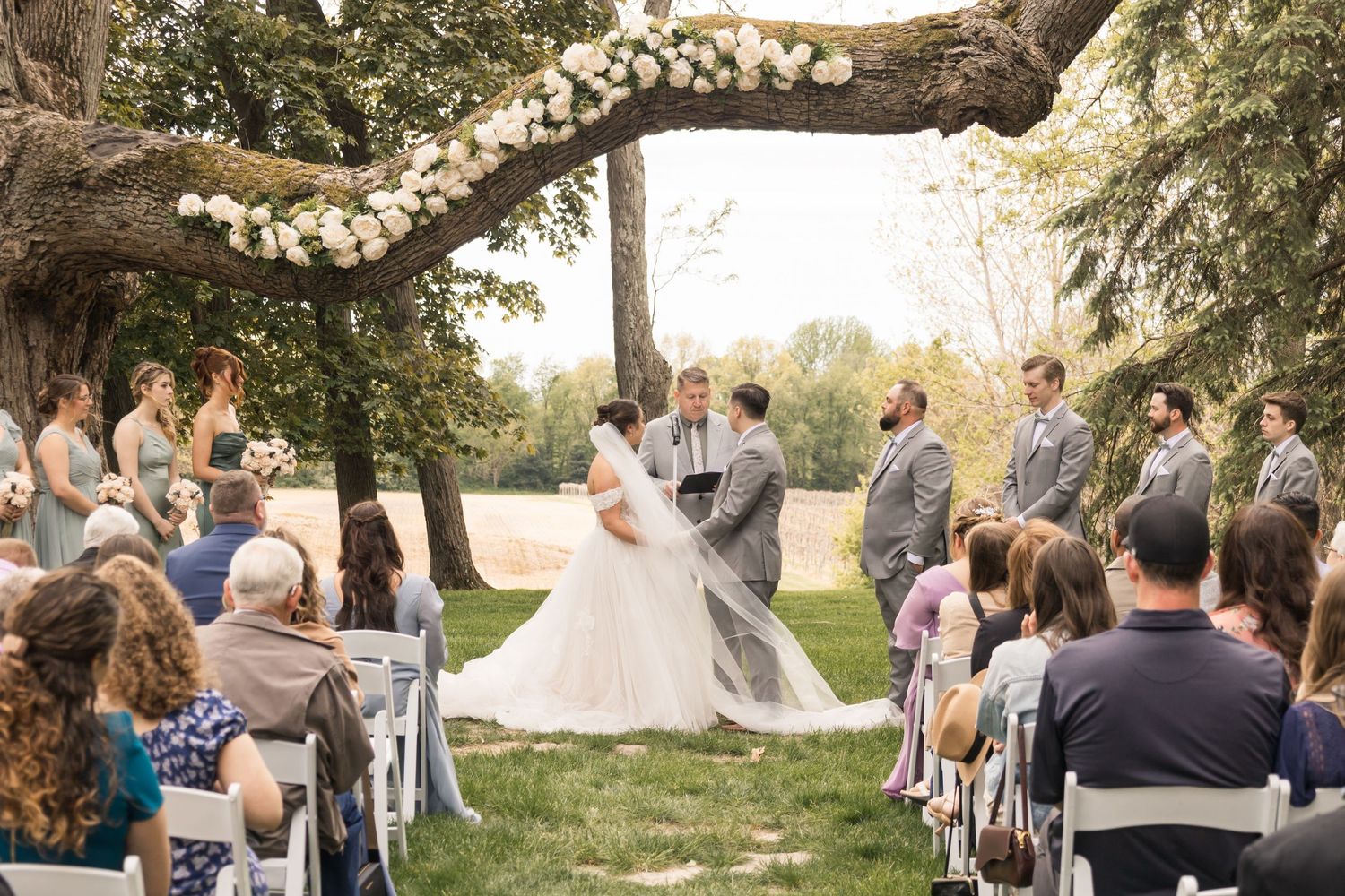 Outdoor wedding ceremony under a flower-adorned tree with bride, groom, and guests seated on white chairs.