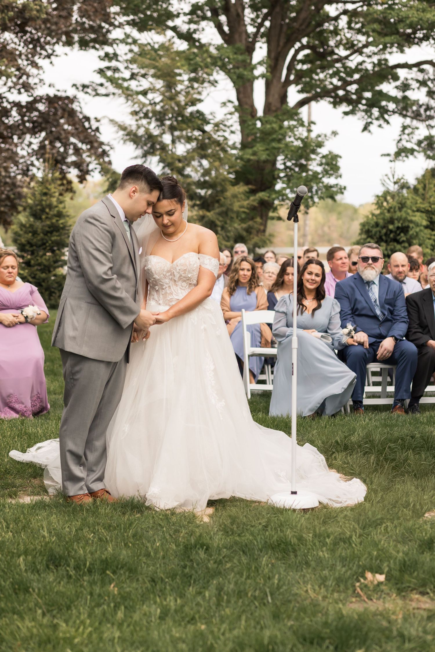 Bride and groom share an intimate moment during outdoor wedding ceremony on green lawn with seated guests in background.