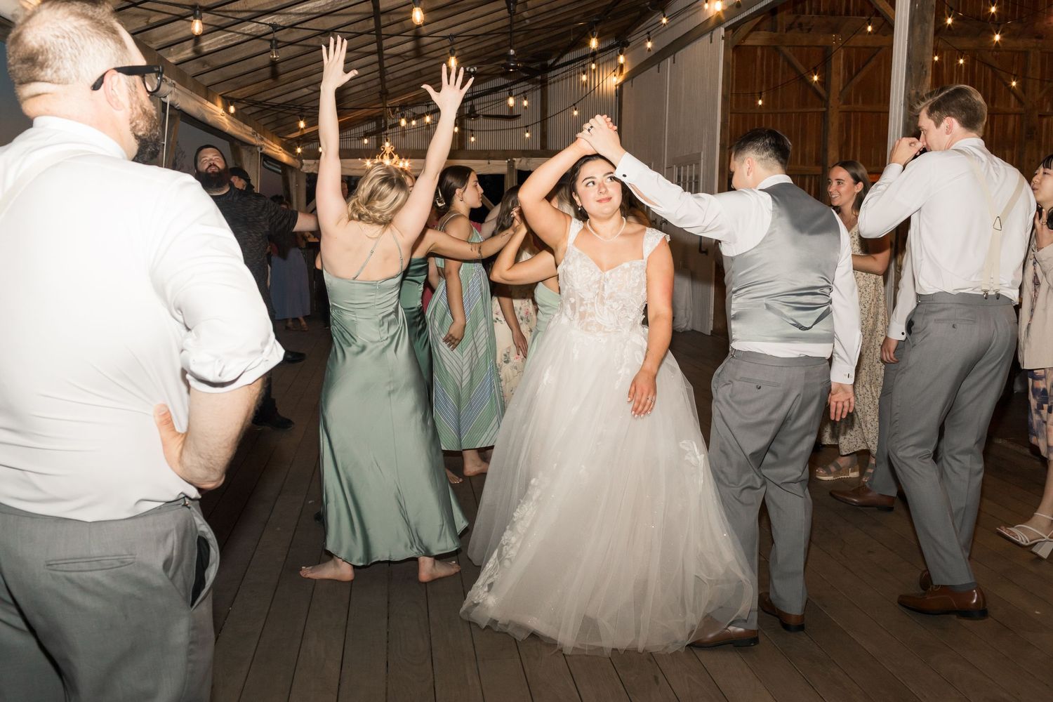 Bride in white tulle gown dancing with guests at wedding reception in rustic barn venue with string lights.