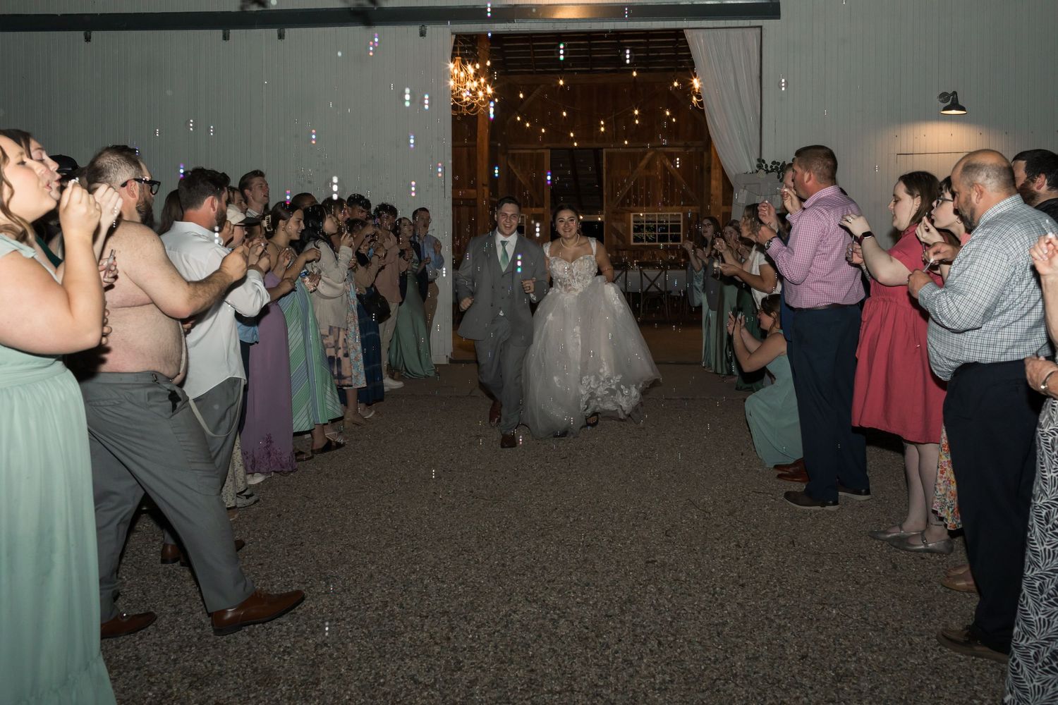 Bride and groom walk through cheering guests at elegant barn wedding reception with string lights.