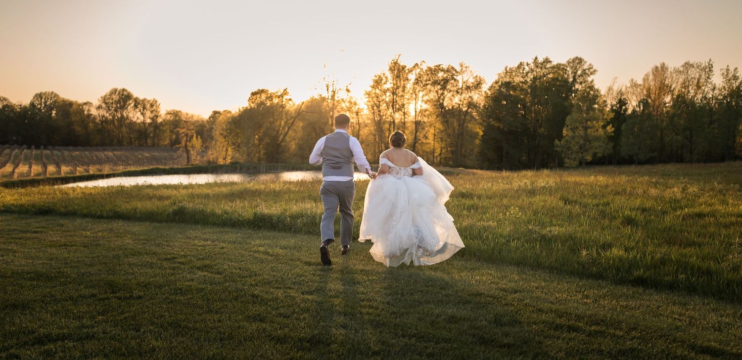 Bride and groom running through a sunlit green field at golden hour with trees and a pond in the background.